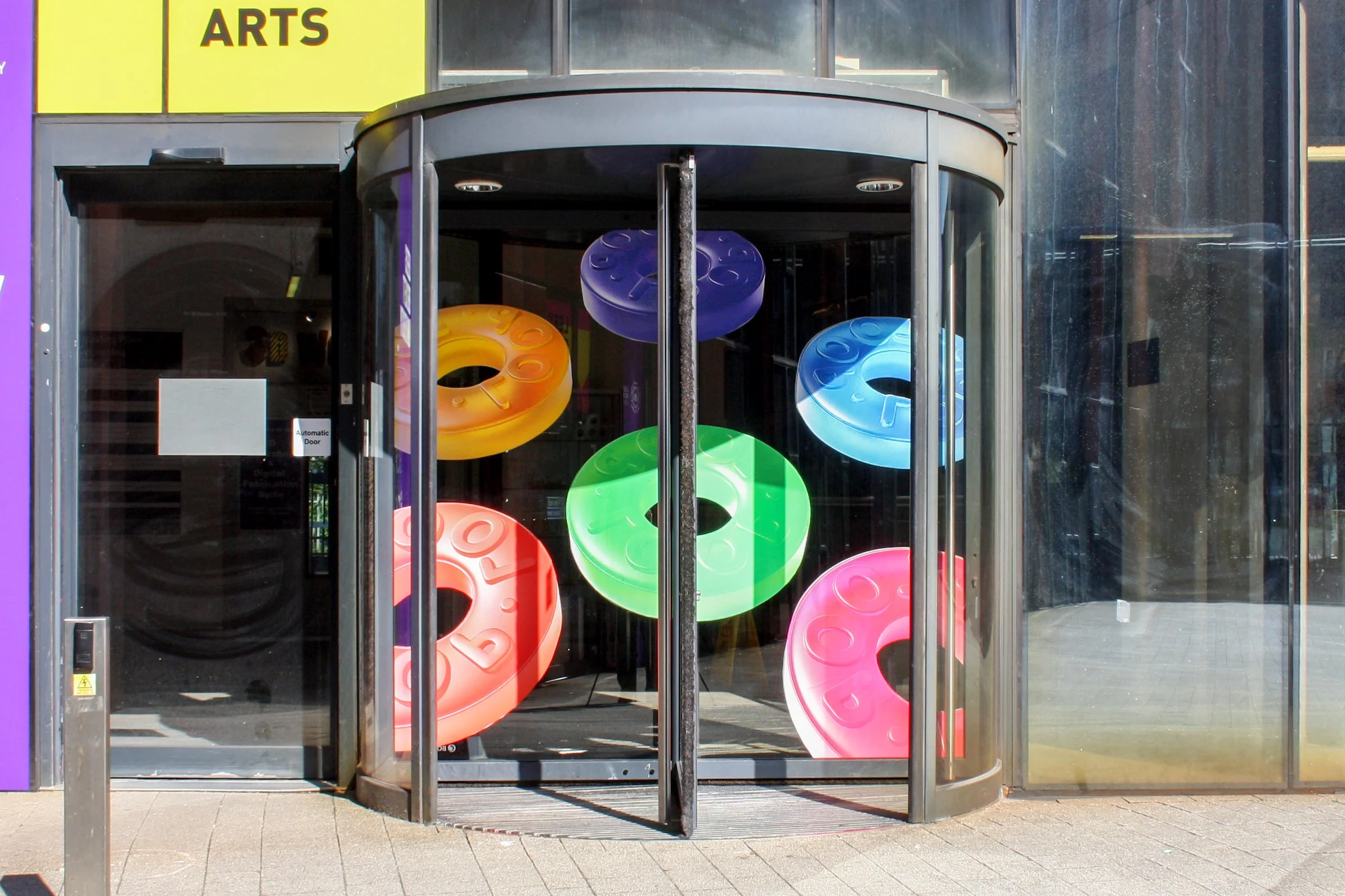 Installation photo of ‘Suite: Repeat’ a vinyl Installation of giant ‘Fruit Loops’ sweets (a variant of a ‘POLO’), with the embossed word ‘LOOP’ instead of ‘POLO’ in the colours pink, blue, green, red, yellow, and purple. Installed on the rotating doo
