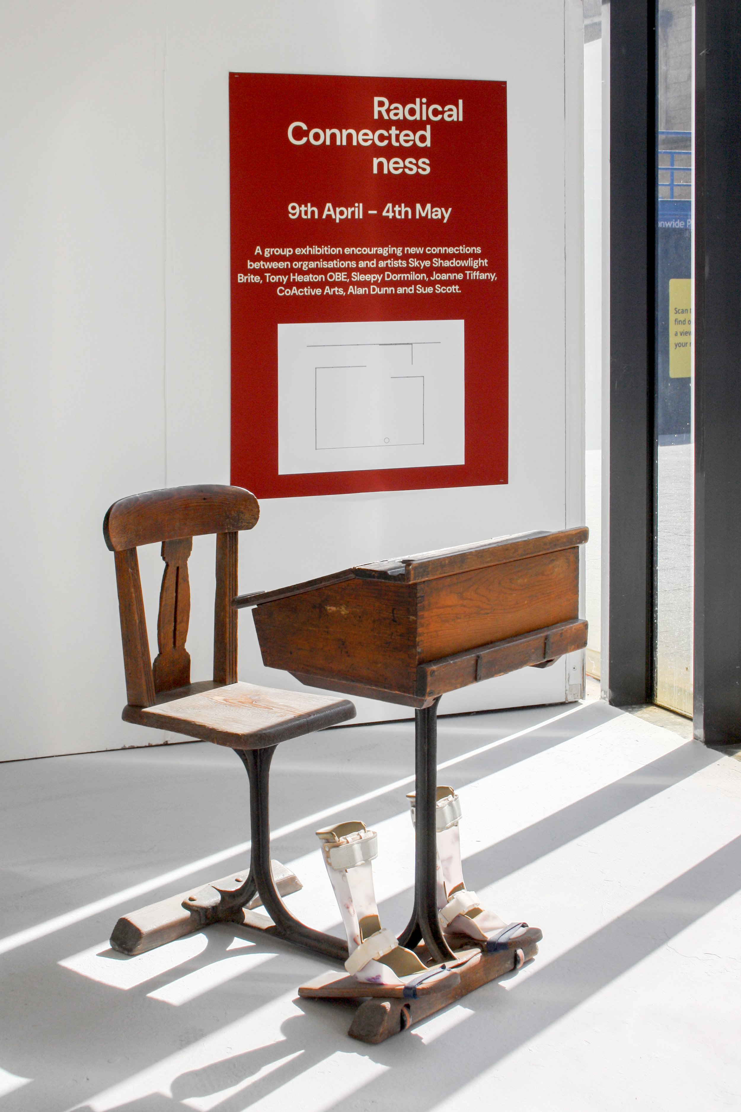 Exhibition photograph of a three-quarter view of the sculptural artwork ‘Brace’ against a white wall and a Dove grey floor. The sculpture is an old worn dark brown wooden Victorian school desk with an attached chair. Underneath is a pair of child-siz