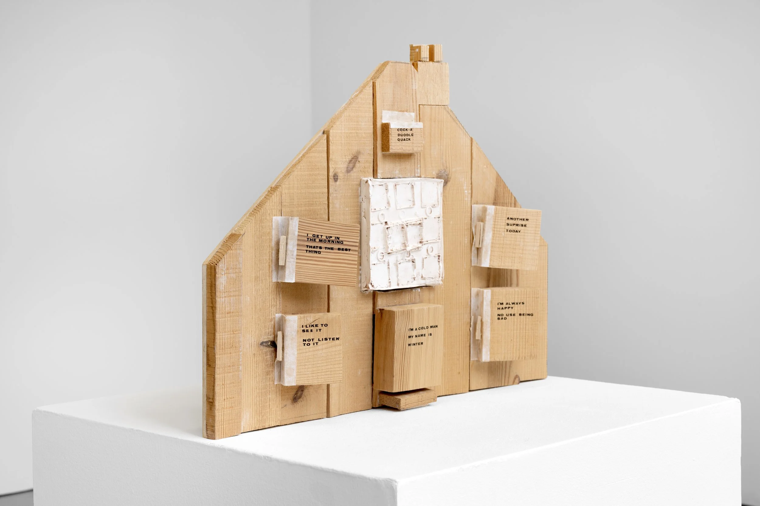 Three quarter exhibition photo of Vivify Project's ‘Wooden Dream Houses’ displayed on a white plinth placed on a grey floor within the White Column space. The sculpture depicts the front of a house, made from rough grained timber, jig sawed pieced to