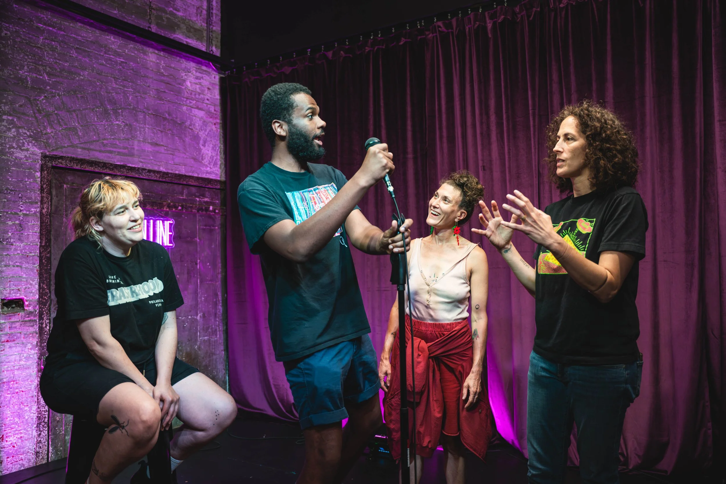 Four people on a stage with purple curtains; one person speaking into a microphone, while others listen and interact.