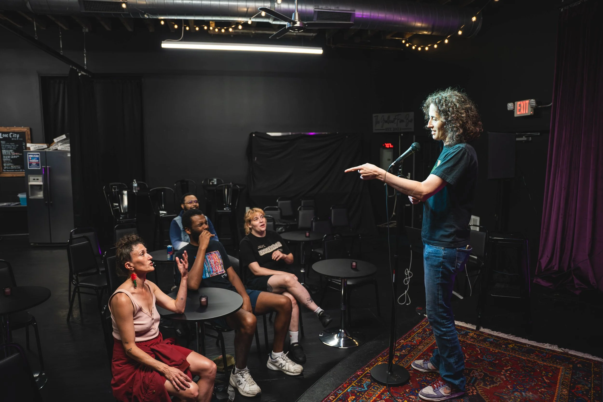 A woman performing stand-up comedy on stage with a microphone, in front of an audience in a small comedy club with black walls and purple curtains.