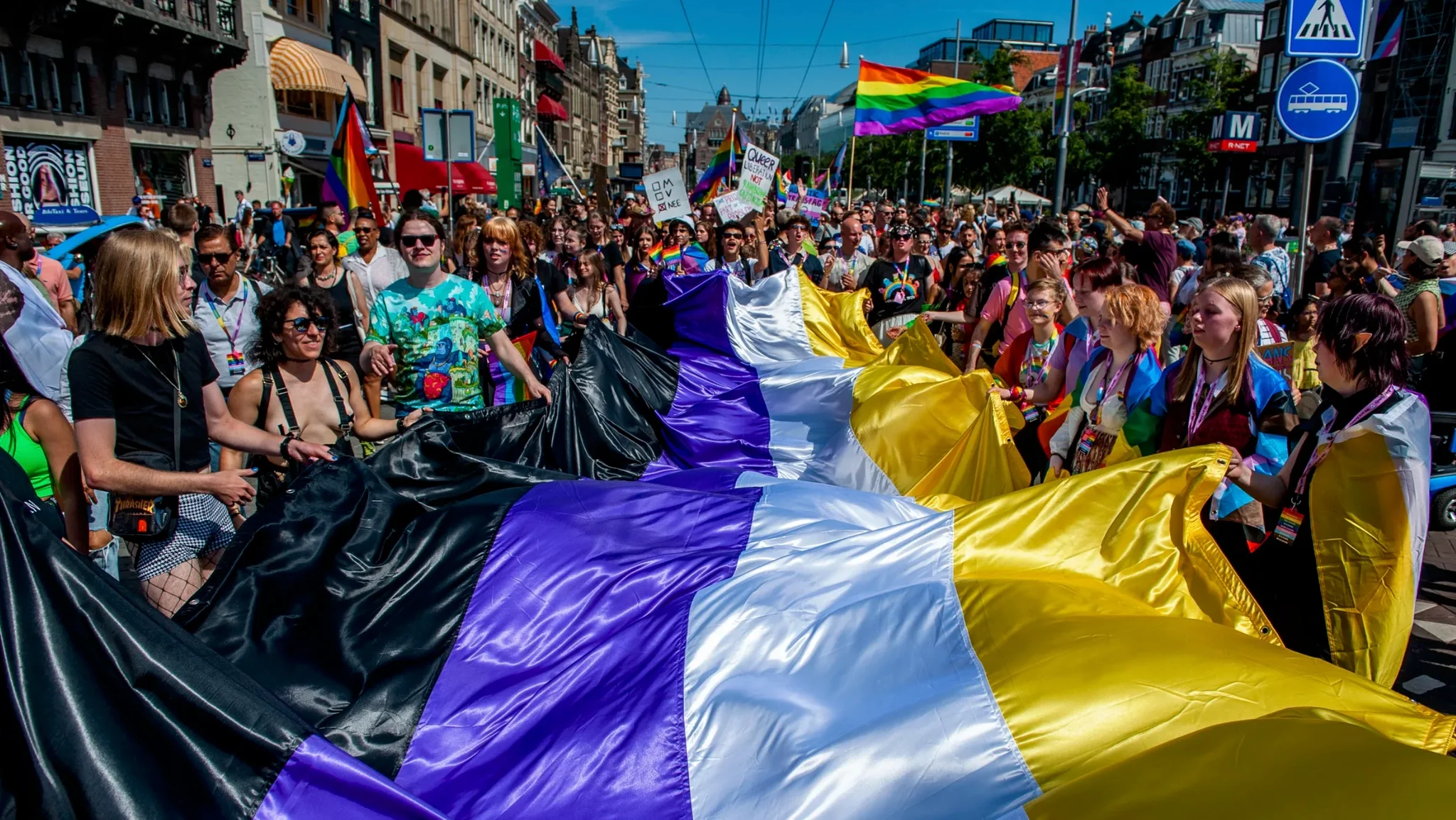 A crowd of people holding a giant non-binary flag at an event that looks like a pride parade