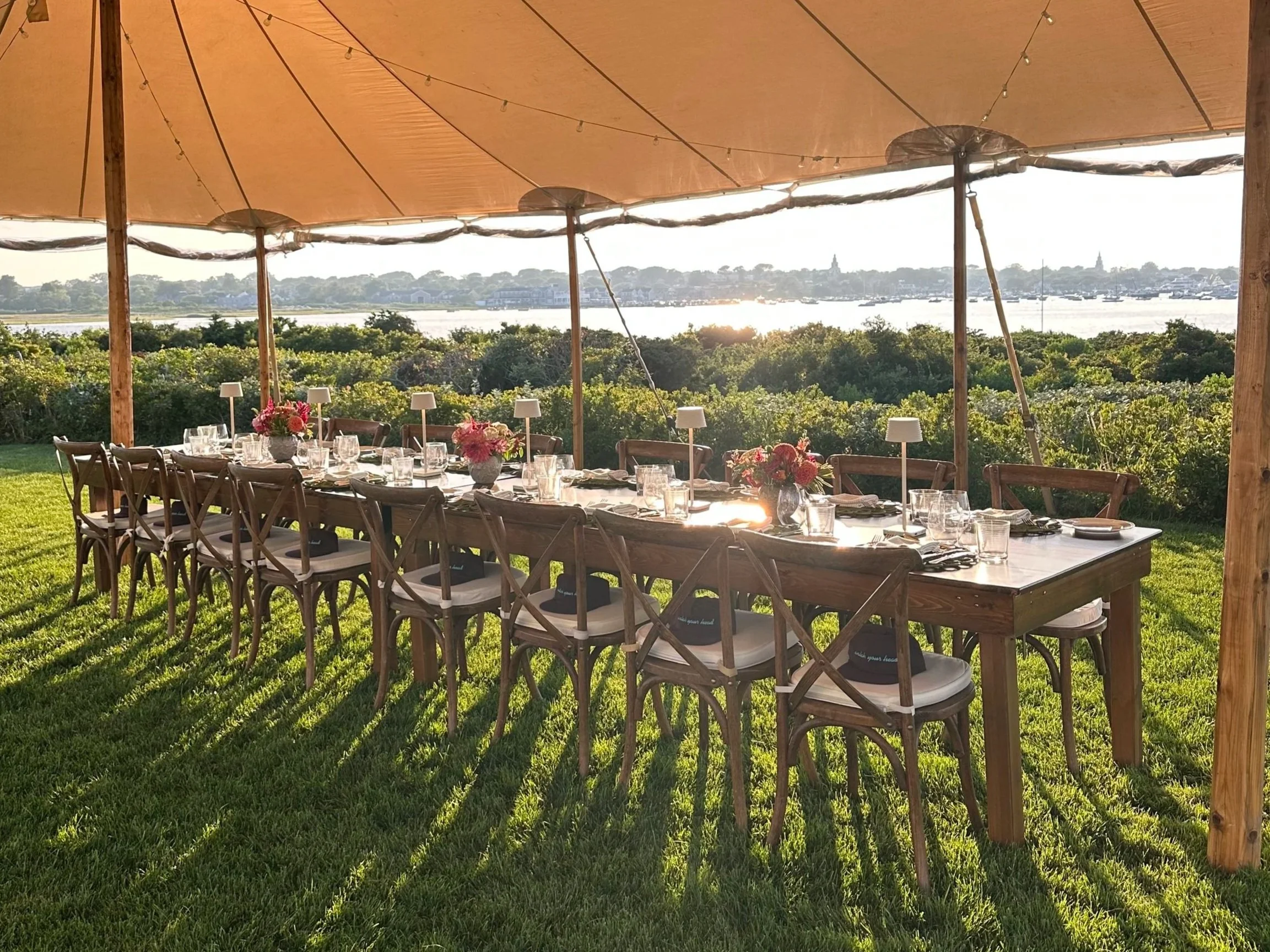 Long outdoor dining table set for a meal catered by Ethos Wine Bar & Bistro in Downtown Nantucket under a beige canopy, with glassware, plates, and pink flowers, overlooking greenery and a the Nantucket Harbor.