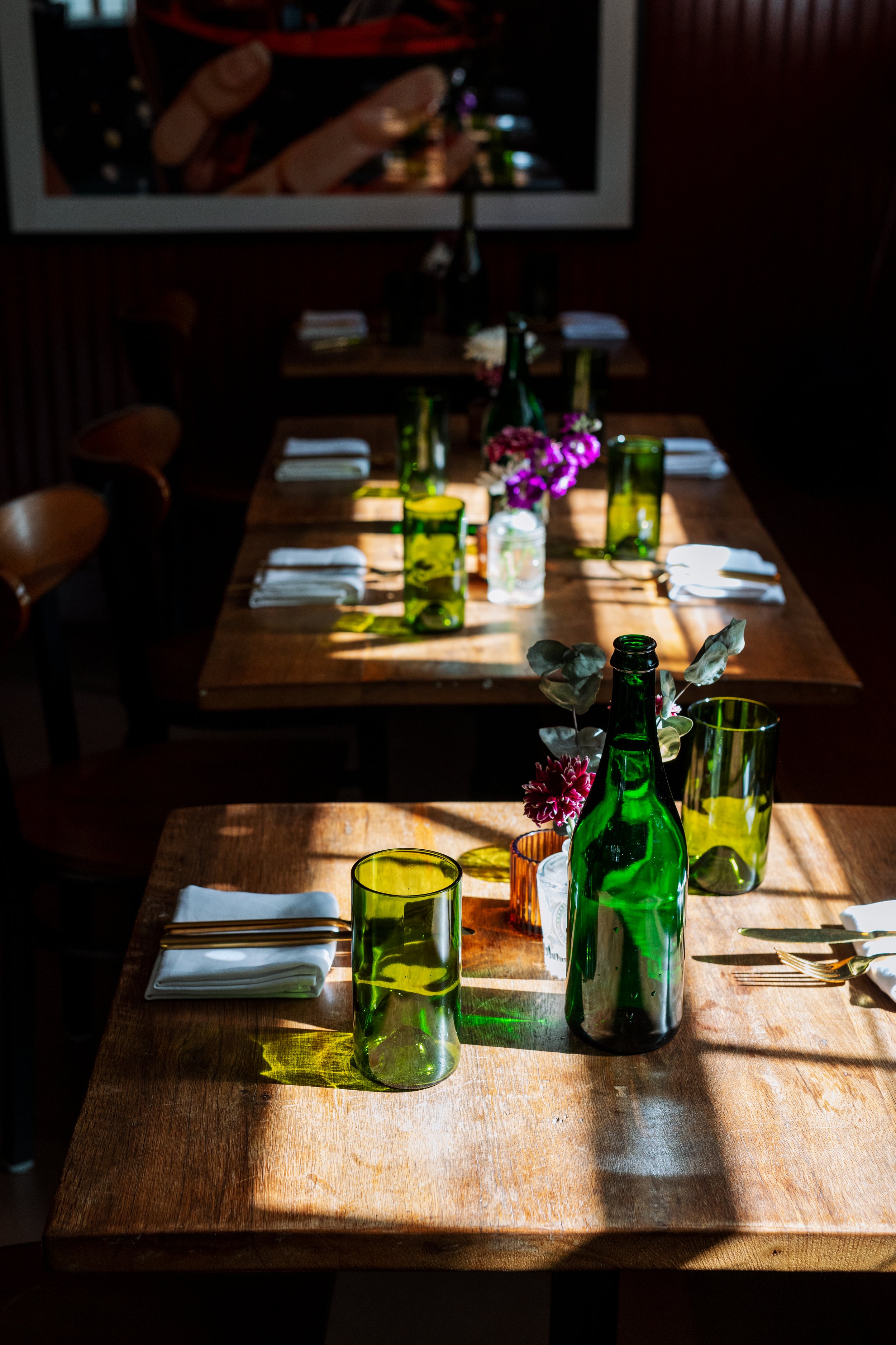 Empty dining tables with green glass bottles, drinking glasses, folded white napkins, silverware, and small flower arrangements, with sunlight casting shadows at Ethos Wine Bar & Bistro in Downtown Nantucket.