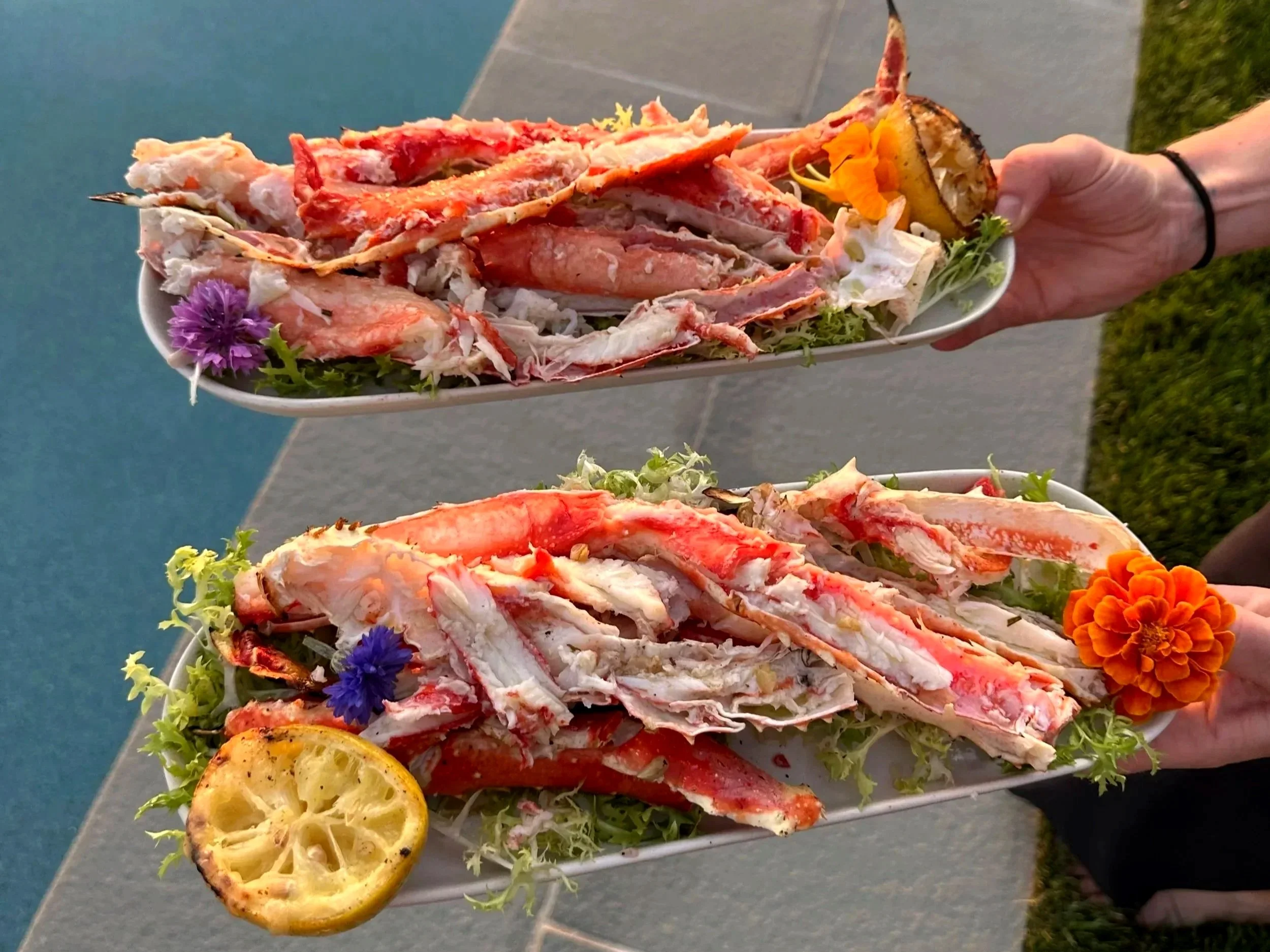 Two white oval plates with king crab legs, garnished with edible flowers, grilled lemon, and herbs, served as a private chef catering event dish by Ethos Wine Bar & Bistro in Downtown Nantucket.