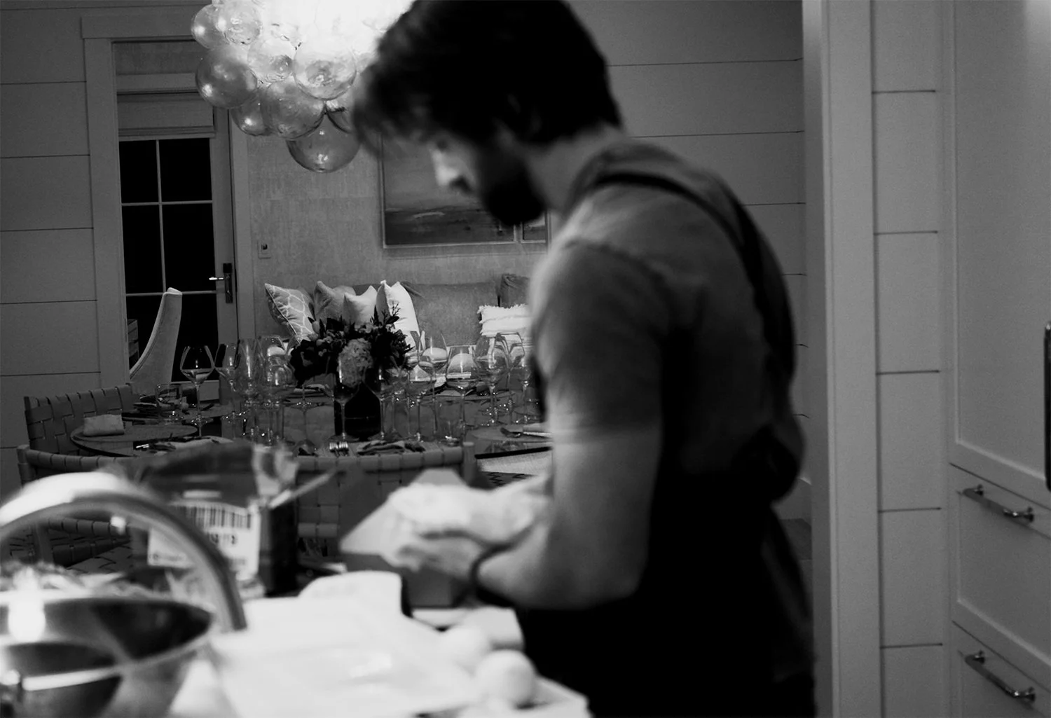 A handsome private chef from Ethos Wine Bar & Bistro in Downtown Nantucket, preparing food in a Nantucket home kitchen. In the background, a dining table with floral centerpiece, glasses, and napkins is set.