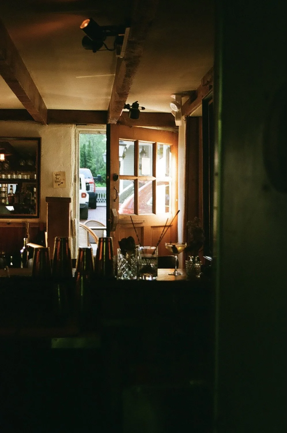Interior of a dimly lit Ethos Wine Bar & Bistro in Downtown Nantucket with wooden beams, a window showing outdoor cars, and a bar counter with cocktail glasses and shakers.