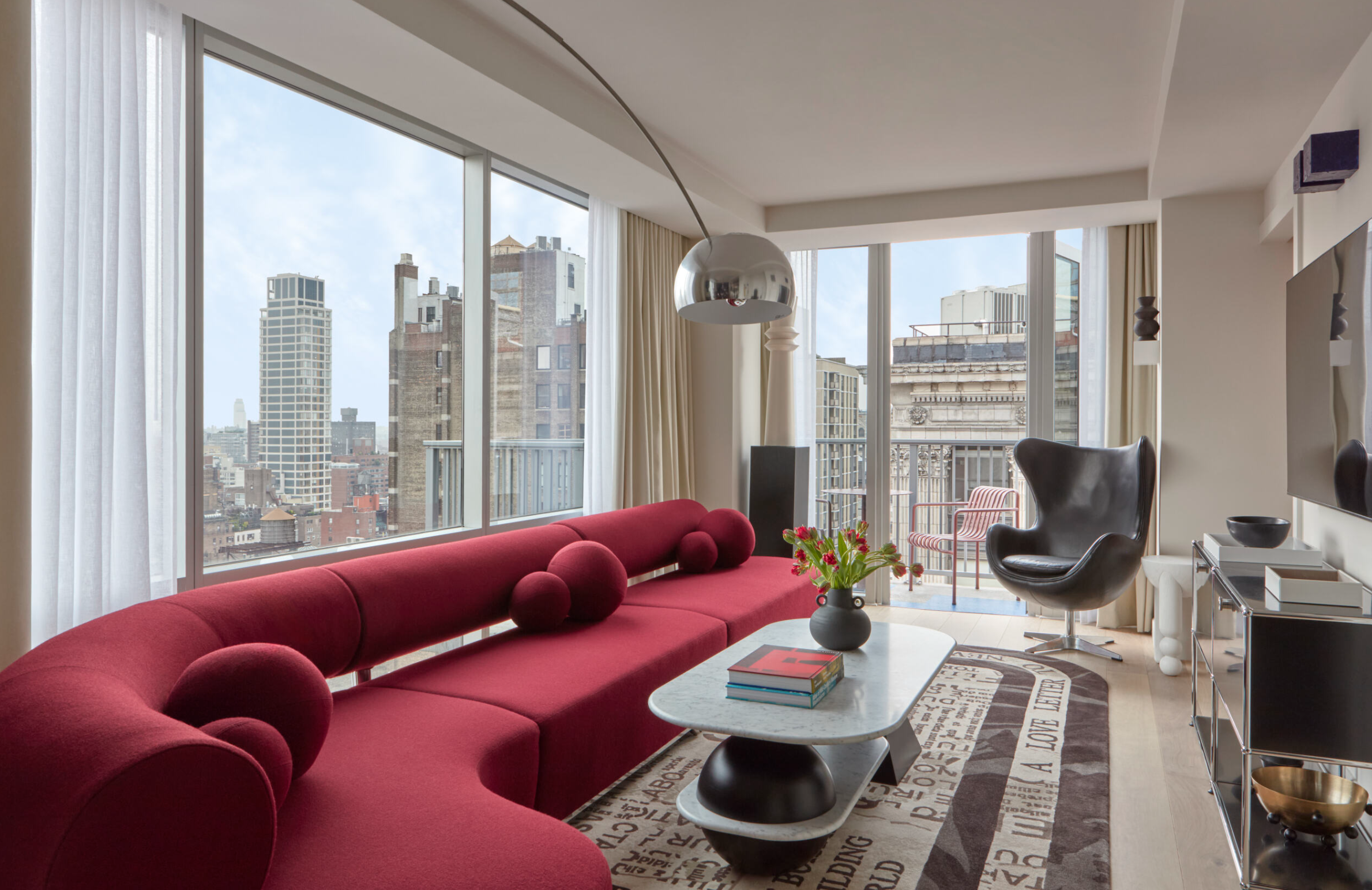 Modern living room with large windows showing city skyline, red sofa, black armchair, white coffee table with books, and decorative items.