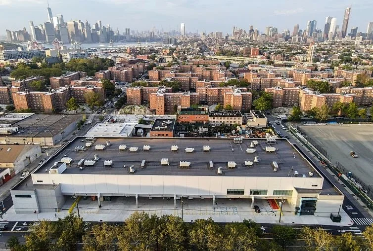 Aerial view of a city skyline with tall buildings in the distance and a large flat-roofed warehouse or industrial building in the foreground.
