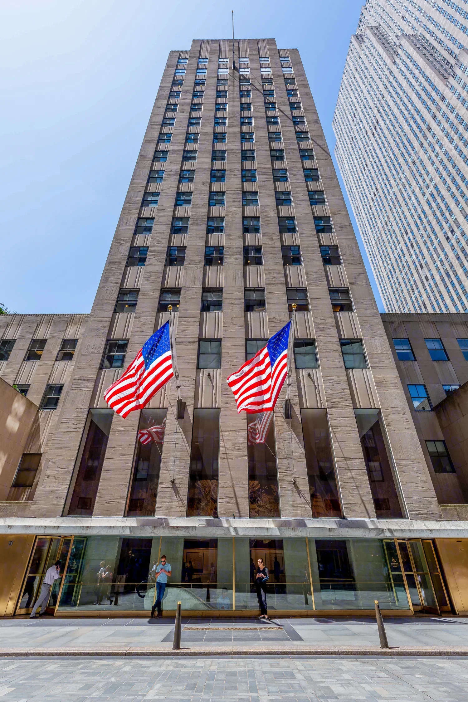 A tall building with many windows and American flags hanging in front, located in an urban area with a glass front entrance and people walking nearby.