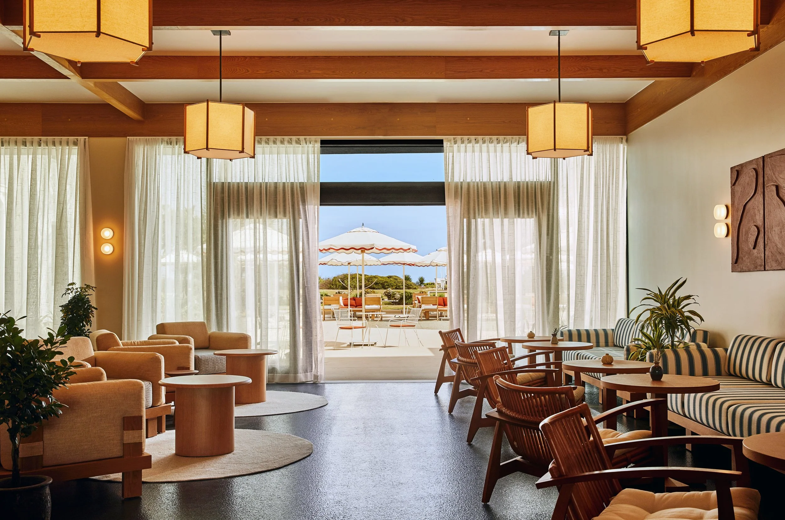 Indoor lounge area with beige and striped chairs, wooden tables, and large windows showing outdoor patio with umbrellas and seating, overlooking the ocean.