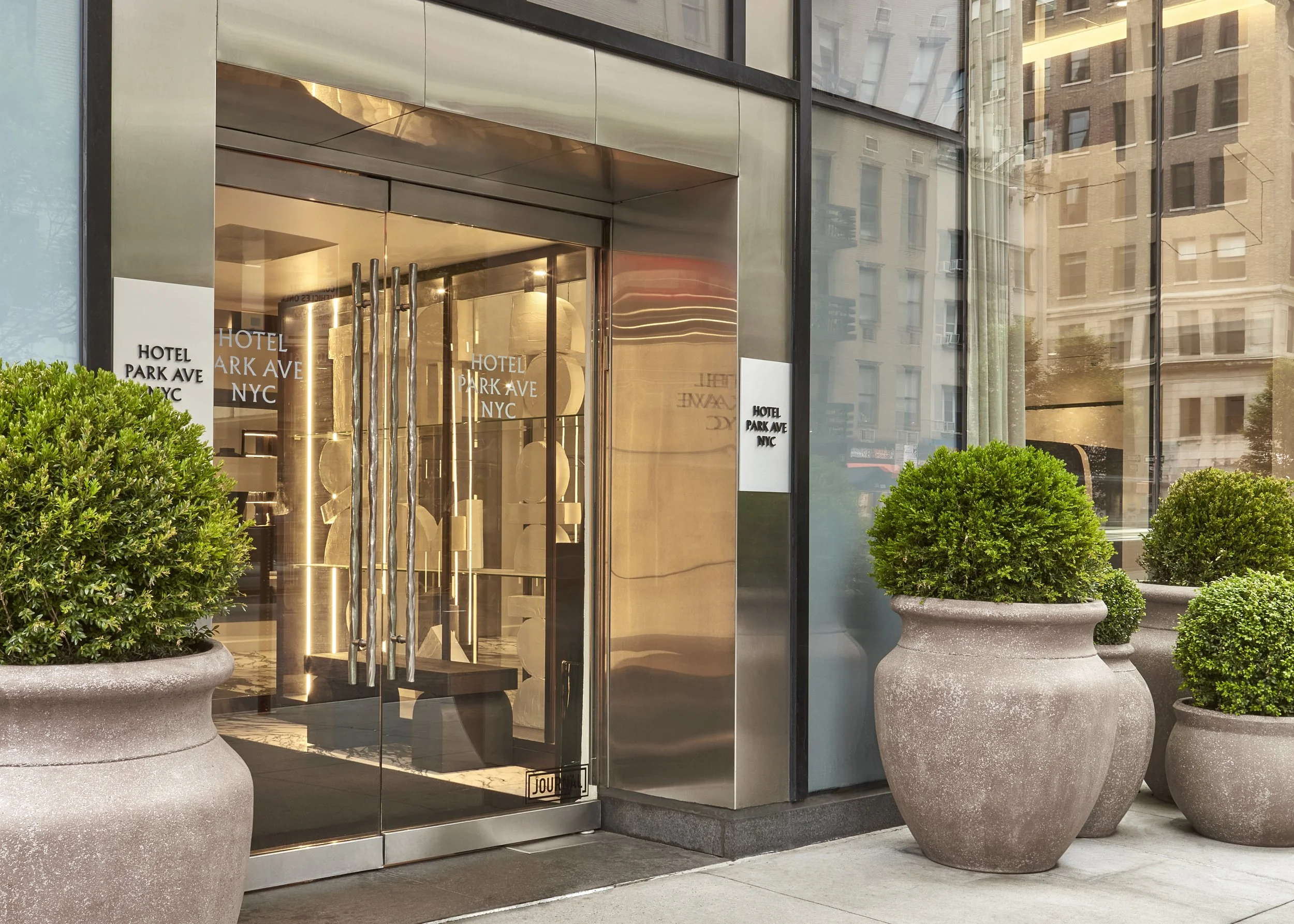 Glass entrance to the Hotel Park Ave NYC with large potted plants on either side.