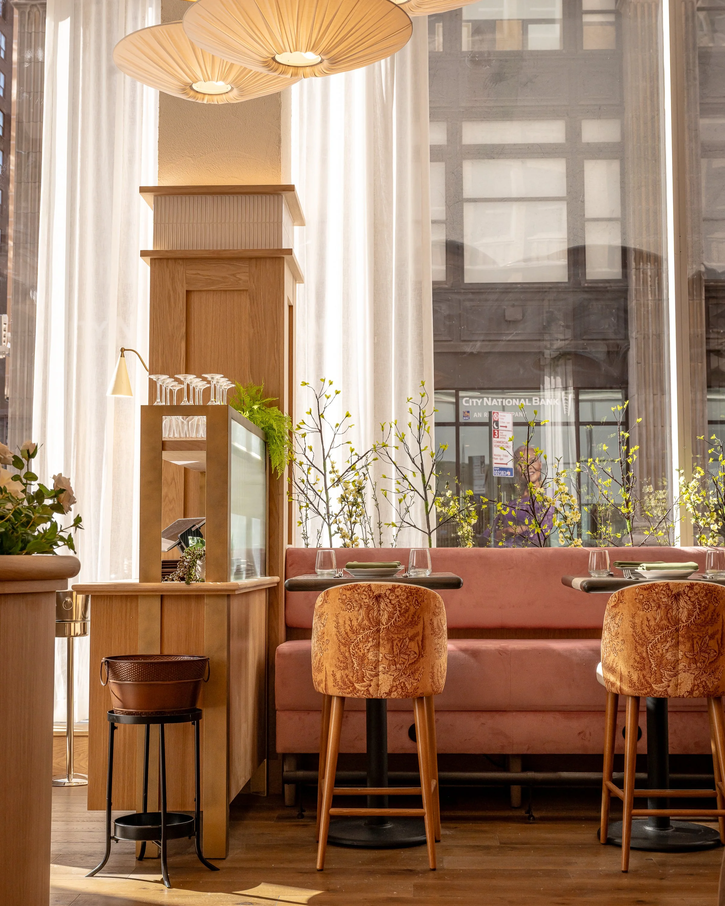 Interior of a restaurant with a view of the outdoor city street through large windows. The space features a high ceiling with wooden pendant lights, a wooden partition, and a cozy seating area with a pink bench, round tables, and upholstered chairs. 