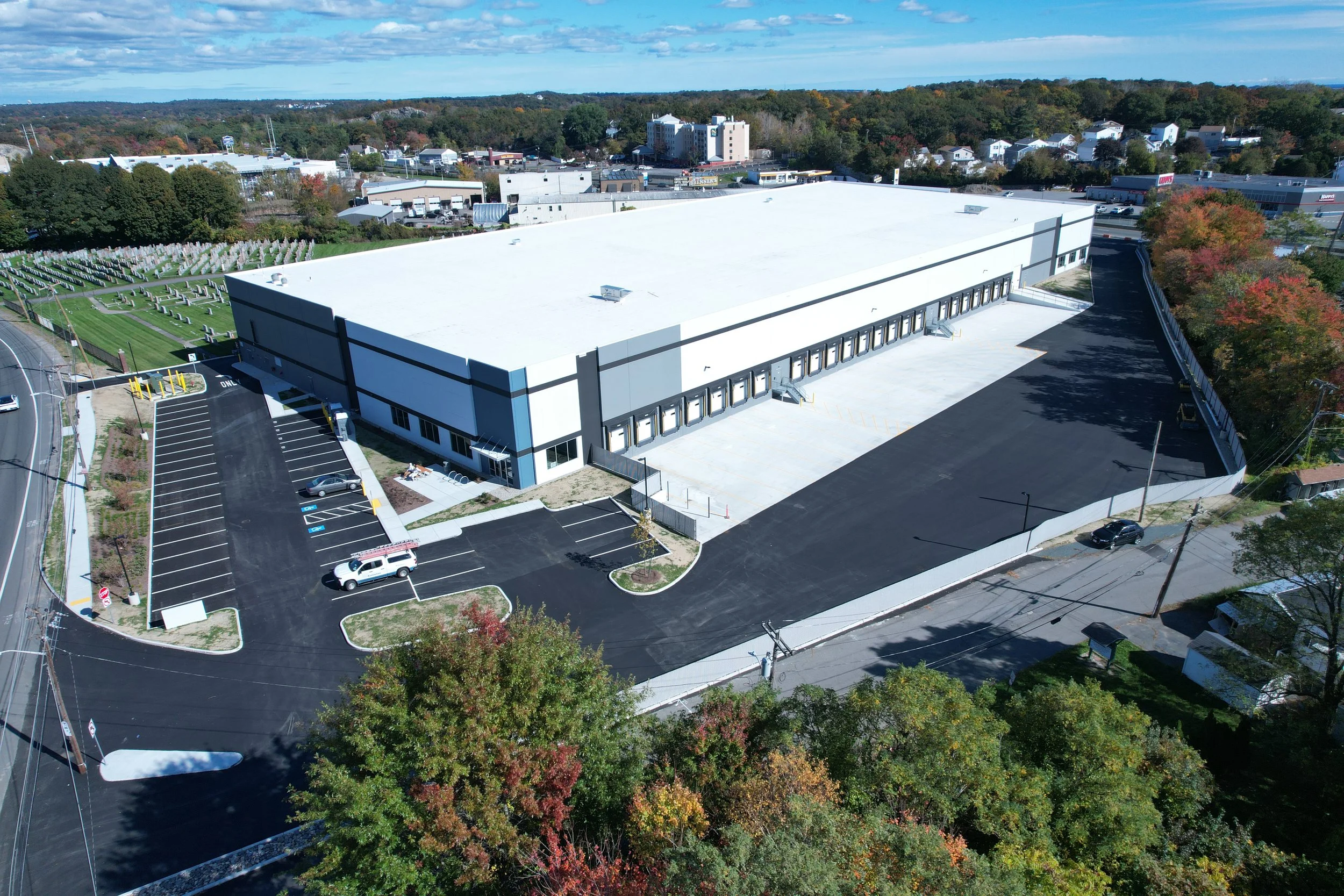 An aerial view of a large white commercial building with surrounding parking lot, trees, and a cemetery with headstones to the left. The background features a small town with commercial and residential buildings and a wooded area with colorful autumn