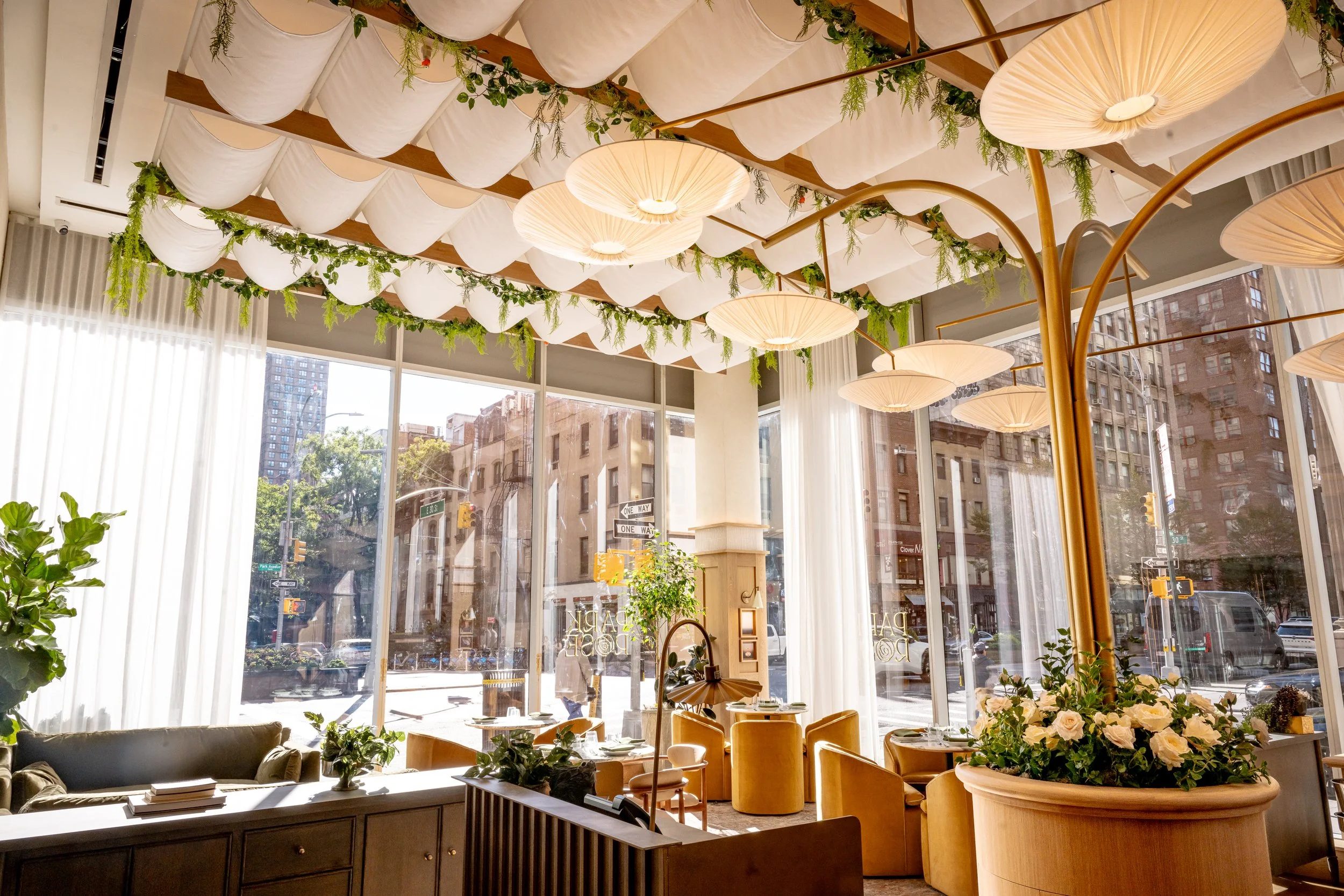 Elegant restaurant interior with large windows, white and gold decor, ceiling drapes, hanging pendant lights, plants, and tables set with tableware and floral arrangements.
