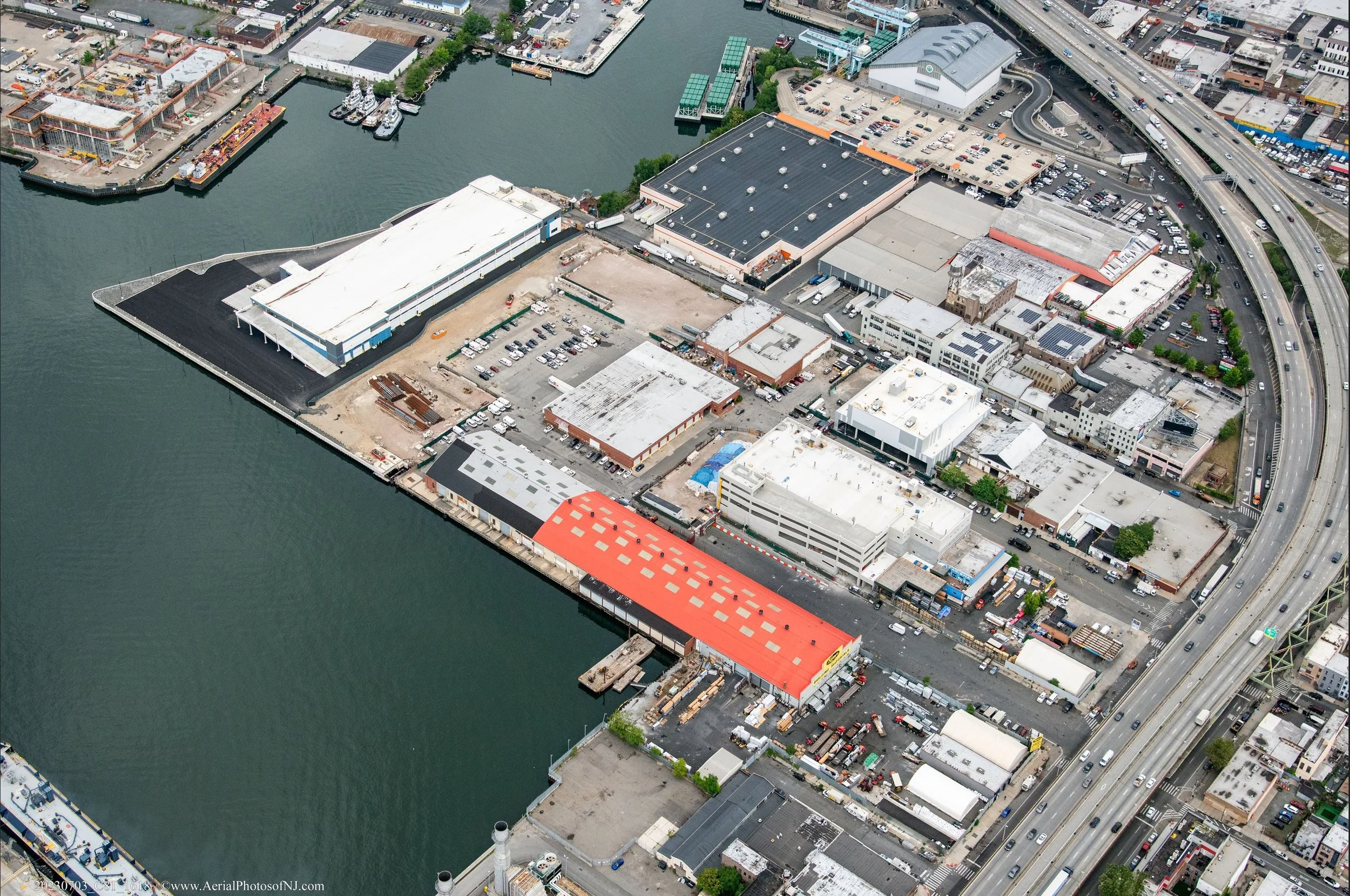 An aerial view of a waterfront industrial and commercial area with warehouses, parking lots, and a marina with boats, alongside a highway with multiple lanes of traffic.