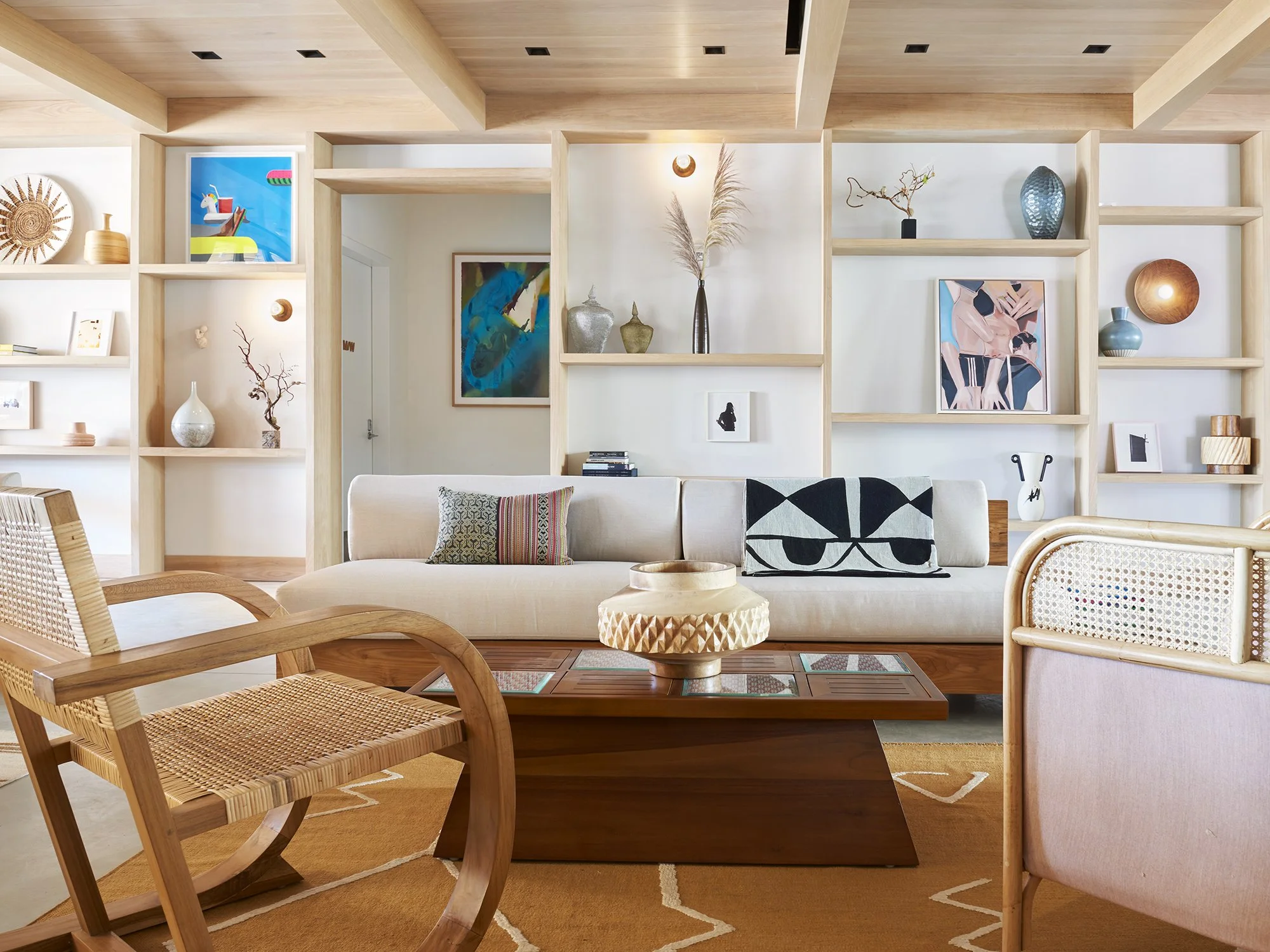 A modern living room with wooden ceiling and walls, decorated with various vases, artwork, and decorative items on open shelves, beige sofa with colorful pillows, and a wooden coffee table with a textured surface. A woven light-colored armchair is in