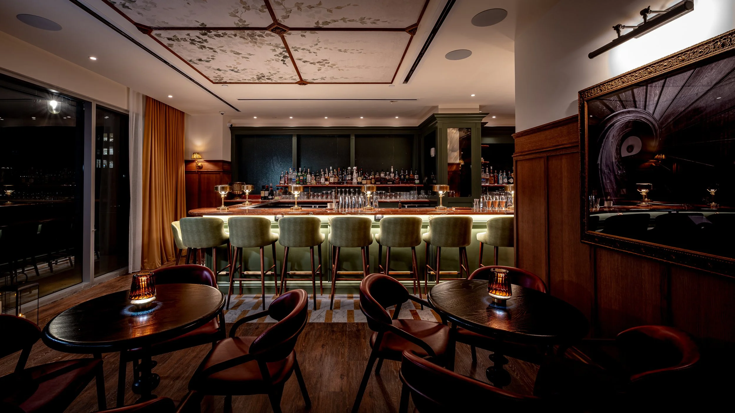 An elegant indoor bar with a marble top counter, bar stools, in a softly lit setting with tables and chairs, large window with curtains, and decorative ceiling and wall features.
