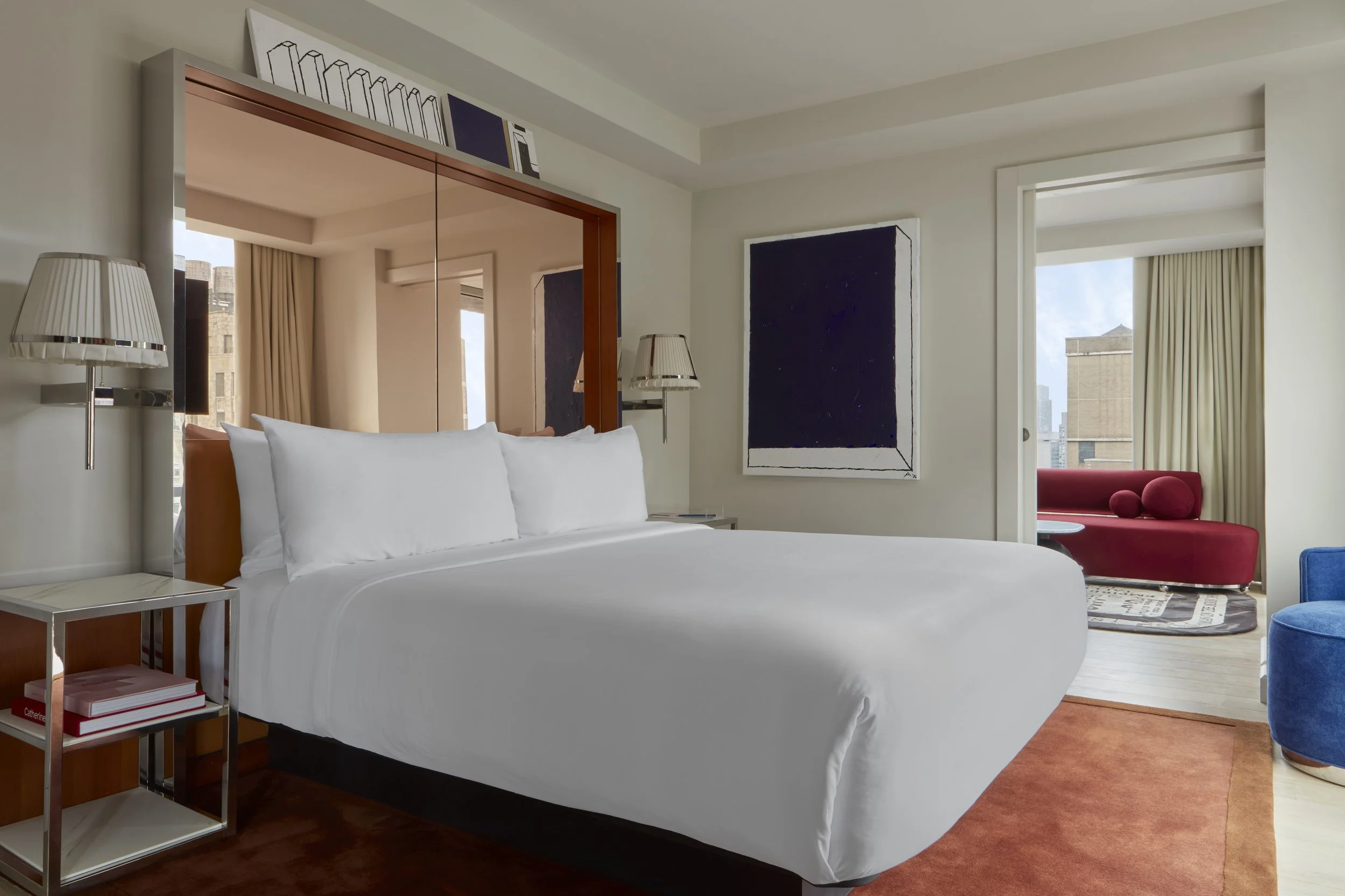 Modern hotel room with a large white bed, mirrored headboard, and wall art, overlooking city buildings through large windows with curtains.