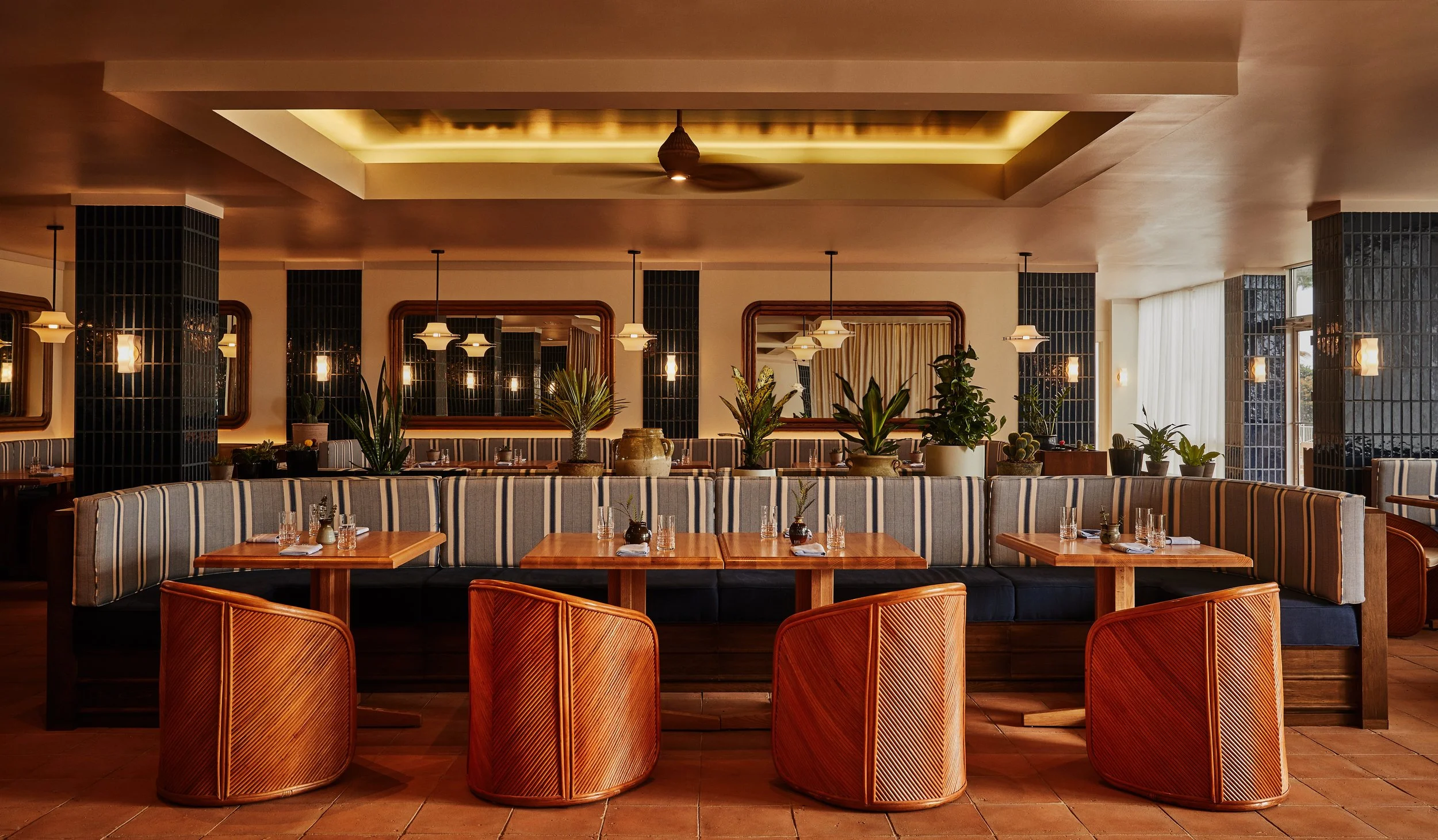 Elegant restaurant interior with striped upholstered booths, wooden tables set with glassware, and potted plants, illuminated by pendant lights and wall sconces.