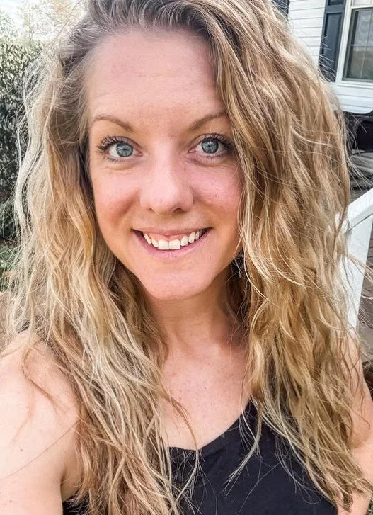 Close-up of a smiling woman with long, curly blonde hair, wearing a black top, outdoors with a house and trees in the background.