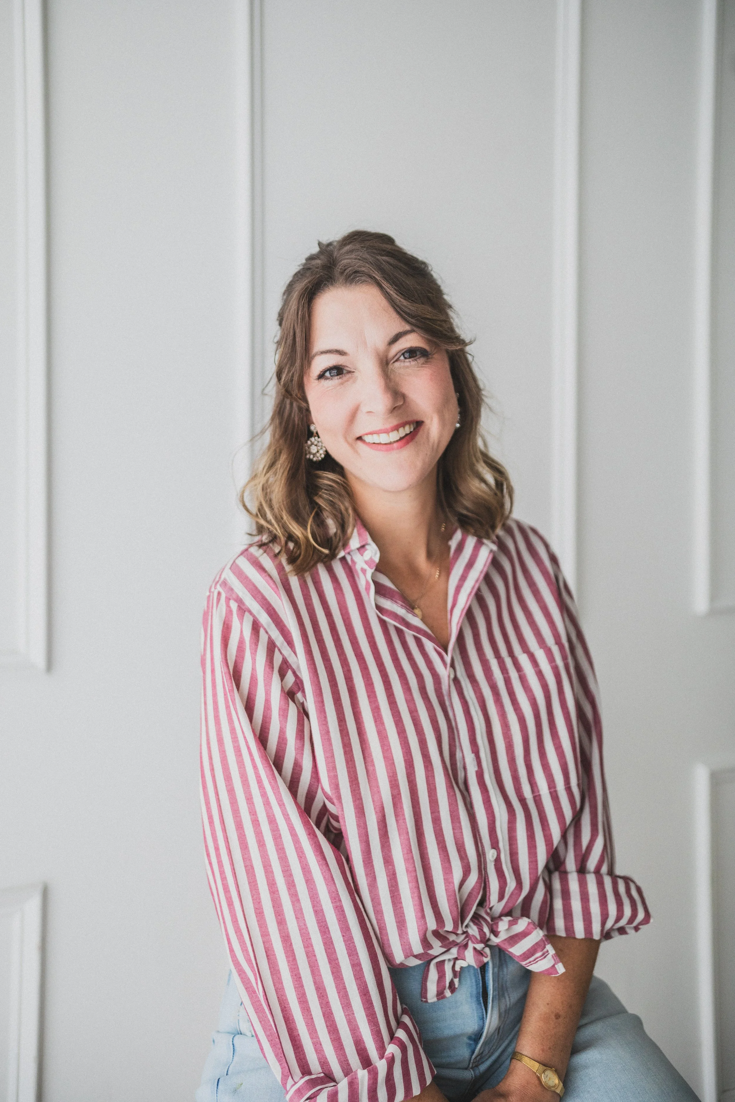 A woman with wavy shoulder-length brown hair smiling, wearing a pink and white striped shirt tied at the waist, light blue jeans, and floral earrings, sitting against a plain white wall.