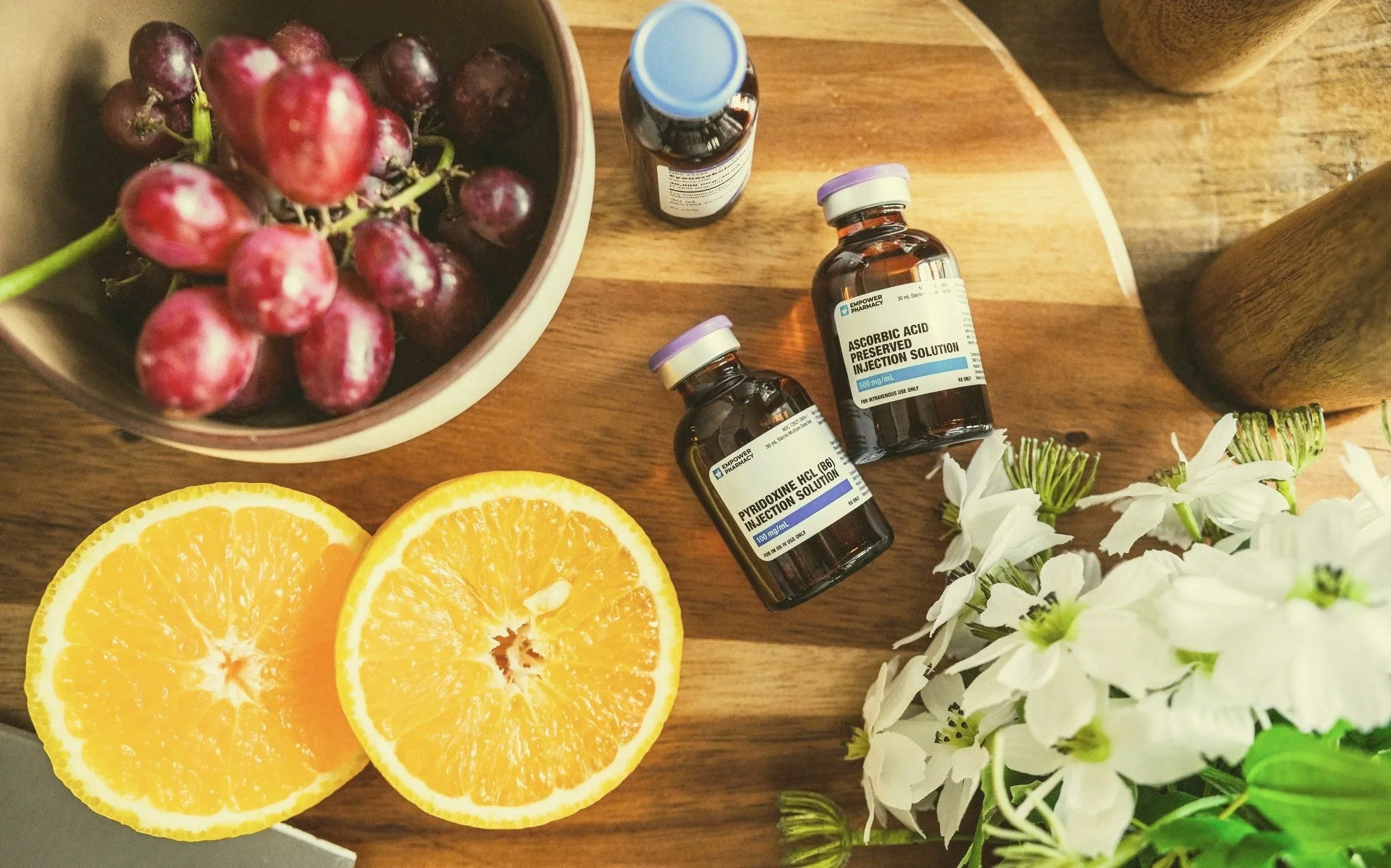 Two halves of an orange, a bunch of red grapes in a bowl, two small medication bottles labeled 'Pyridoxine HCL' and 'Ascorbic Acid', and white flowers on a wooden surface.
