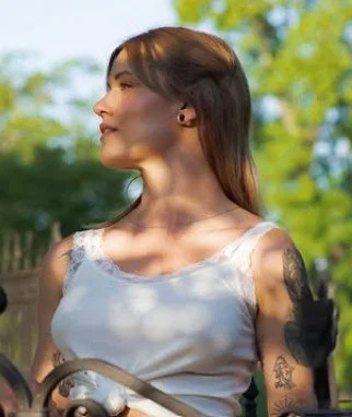 A woman with long brown hair, tattoos on her arms, and wearing a white tank top standing outdoors with green trees in the background.
