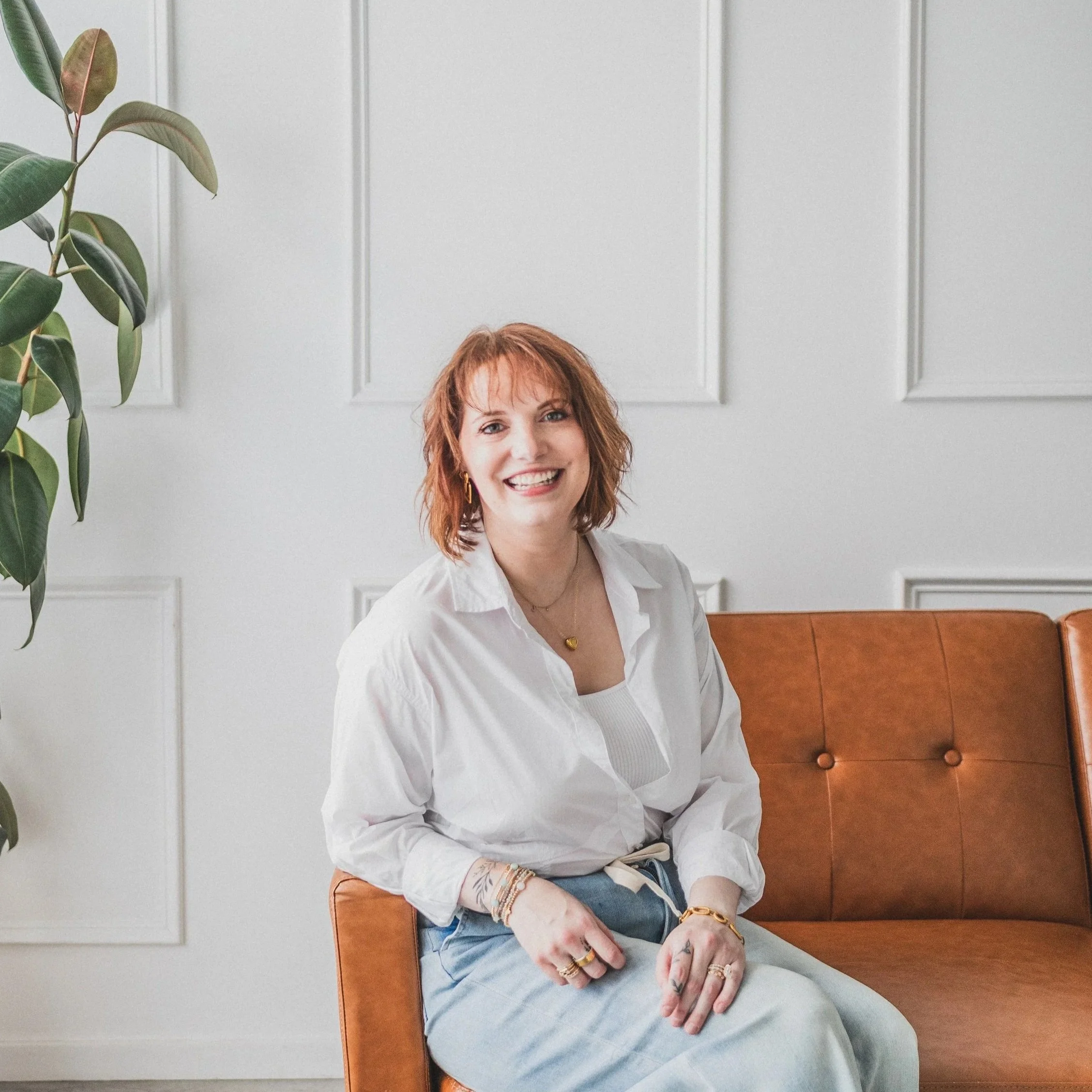 A woman with short red hair sitting on a brown leather chair in a bright, white room with decorative white wall paneling, smiling at the camera.