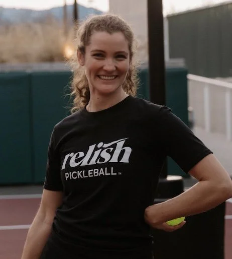 A woman with curly brown hair, wearing a blue T-shirt that reads 'relish PICKLEBALL.', standing against a green wall and smiling.