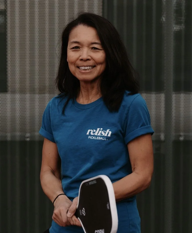 A woman smiling, wearing a blue Relish Pickleball shirt, a black Adidas cap, standing in front of a green background.