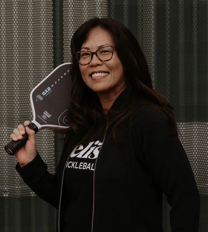 Woman with glasses smiling on a tennis court during sunset.
