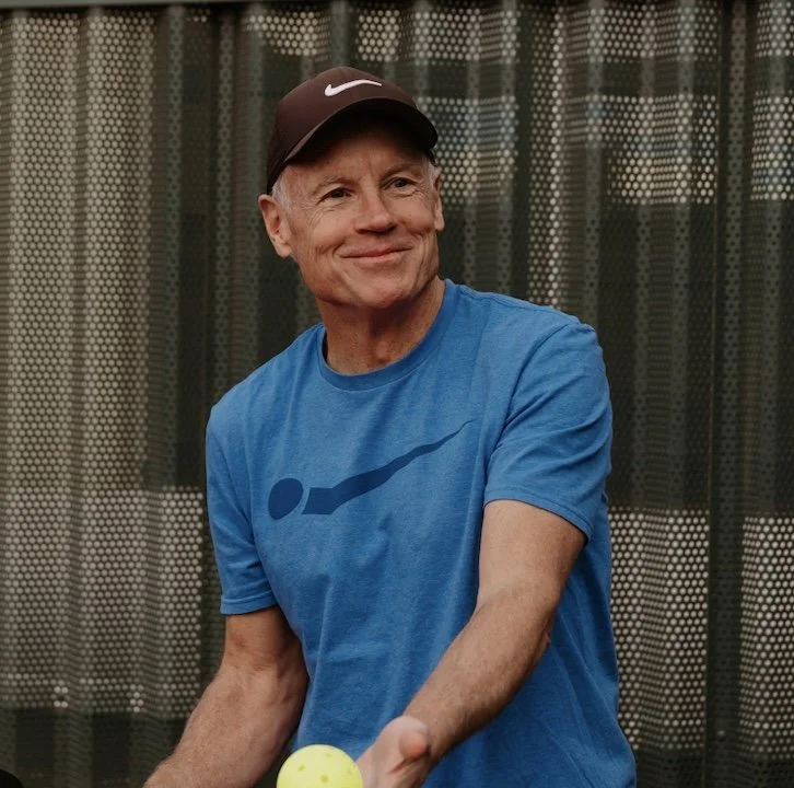 Older man with gray hair smiling, wearing a black 'relish PICKLEBALL' t-shirt, standing against a green background.