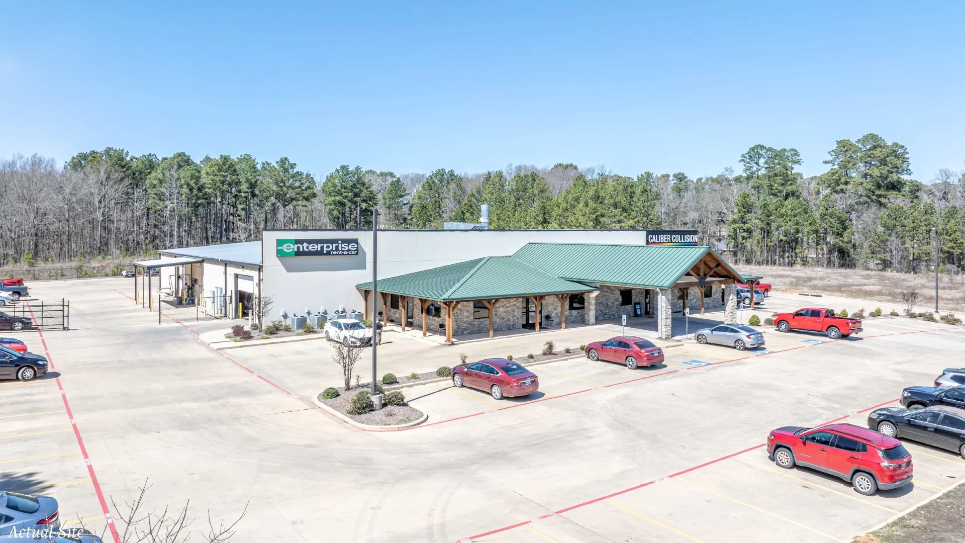 Large parking lot in front of a commercial building with signs for Enterprise Rent-A-Car and Caliber Collision, few cars parked, surrounded by trees.