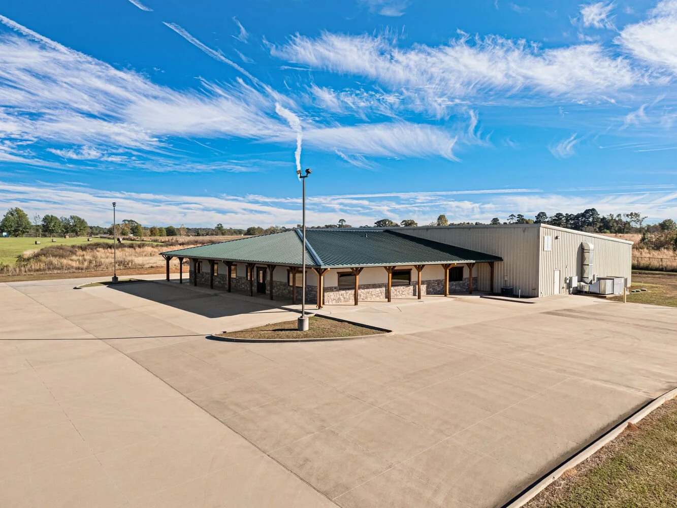 Empty concrete parking lot with a single building, a green metal roof, stone wall accents, and metal exterior walls, under a blue sky with wispy clouds.