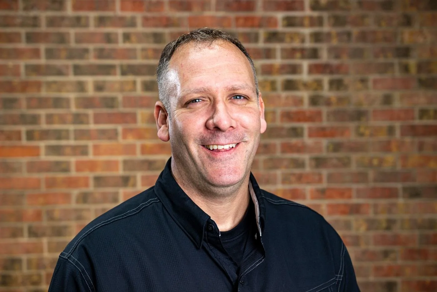 A portrait of a smiling middle-aged man with short hair, wearing a black button-up shirt, standing in front of a brick wall.