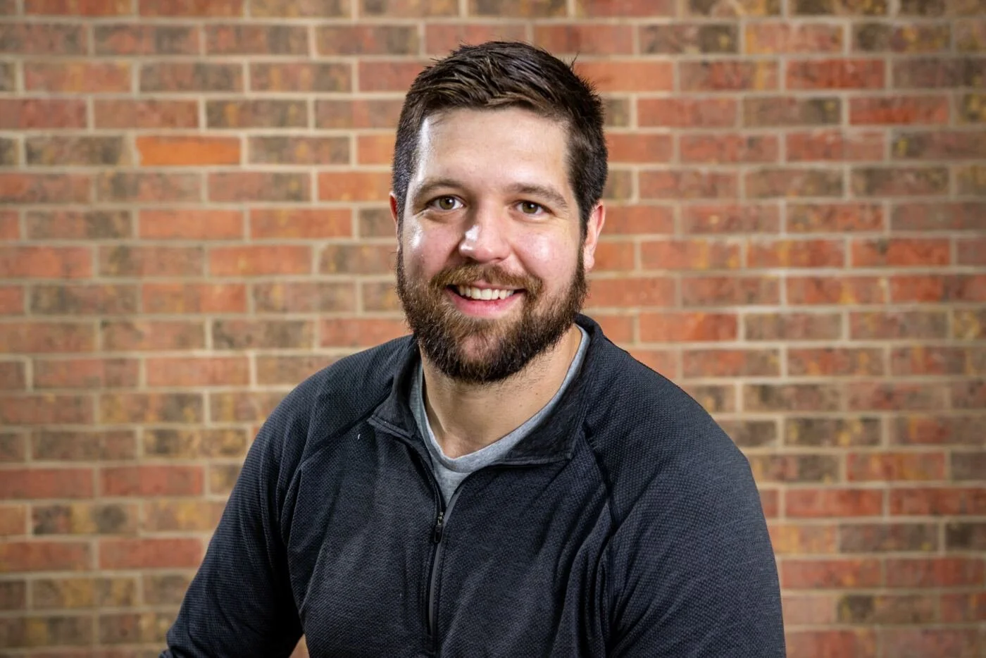 A smiling man with a beard and short brown hair, wearing a black zip-up athletic jacket and a gray shirt, sitting in front of a brick wall.