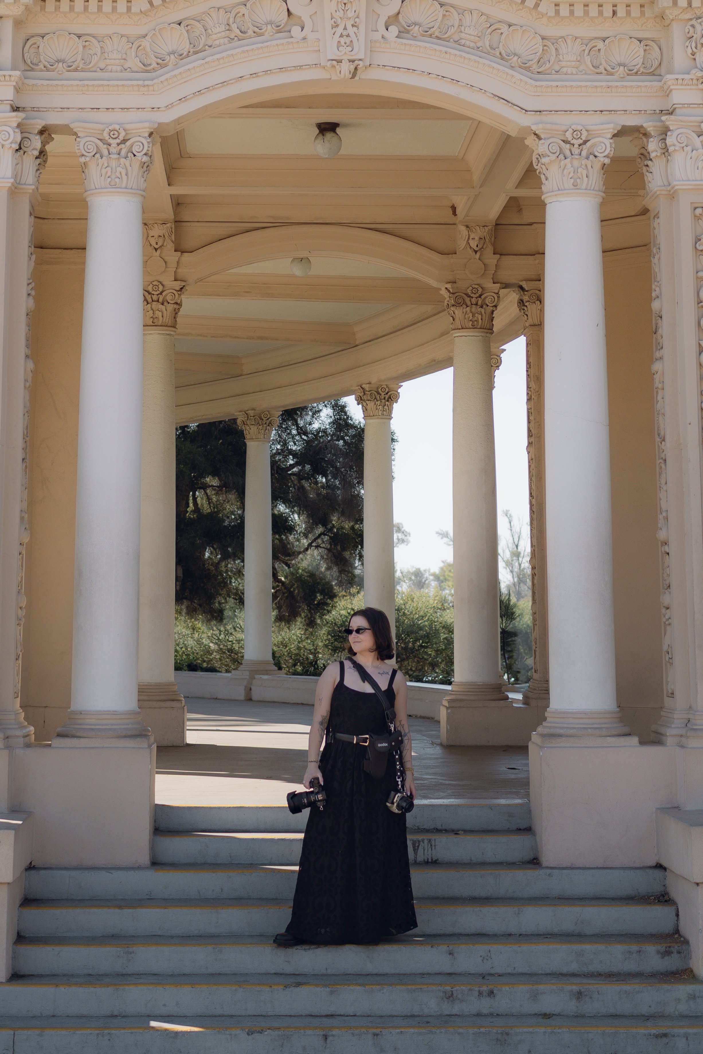A woman in a black dress and sunglasses standing on steps in Balboa Park in San Diego, holding two cameras.