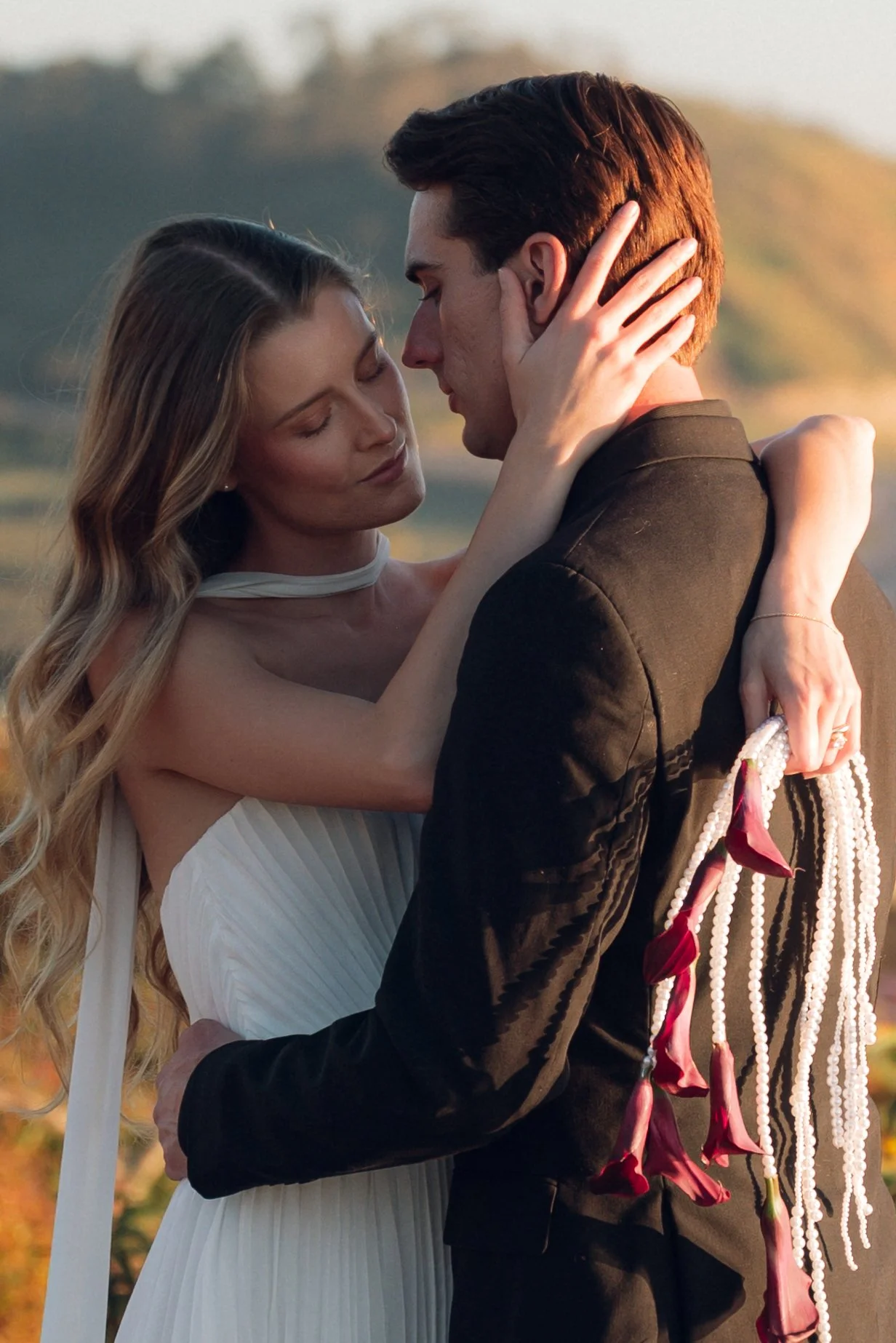 A couple in wedding attire sharing an intimate moment outdoors at sunset, with the woman holding a bouquet of red calla lilies.