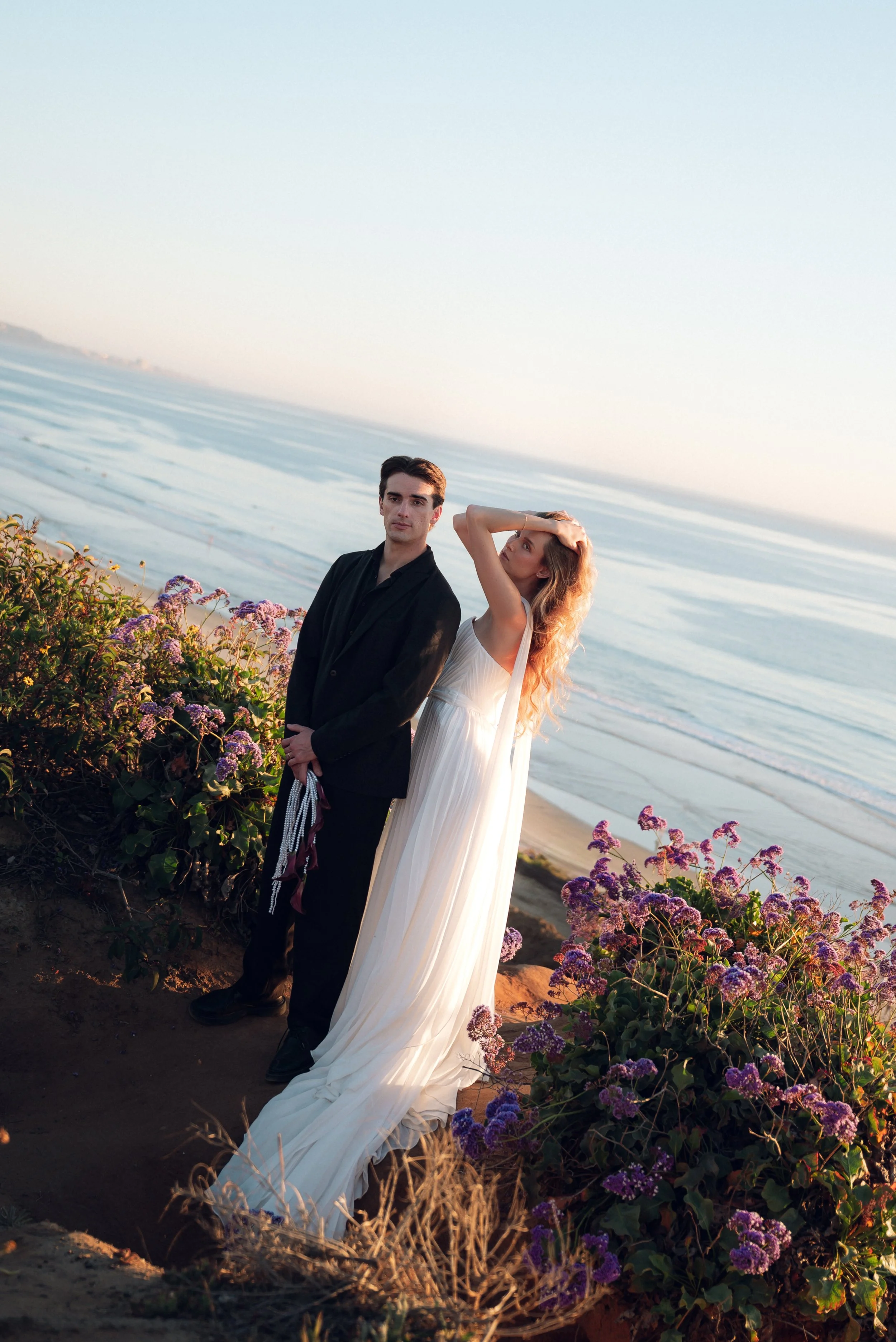 A couple stands at the beach during sunset, with the woman in a long white dress and the man in a black suit, surrounded by purple flowers.