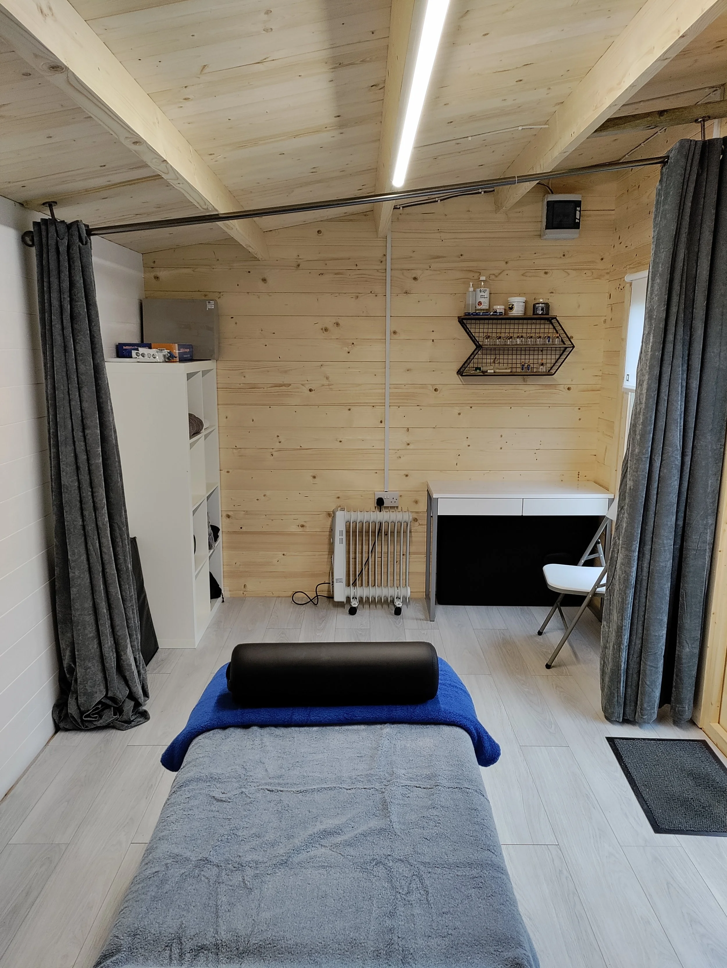 Interior of a small, wooden-paneled room with a massage table, black bolster pillow, gray curtains, shelving, radiator, desk, folding chair, and lighting.