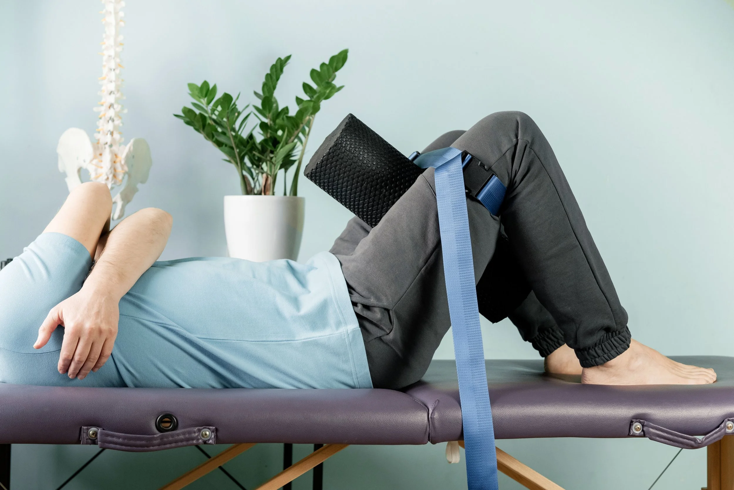 Person lying on a massage table with a foam roller and yoga strap, next to a potted plant and a model of a spine.