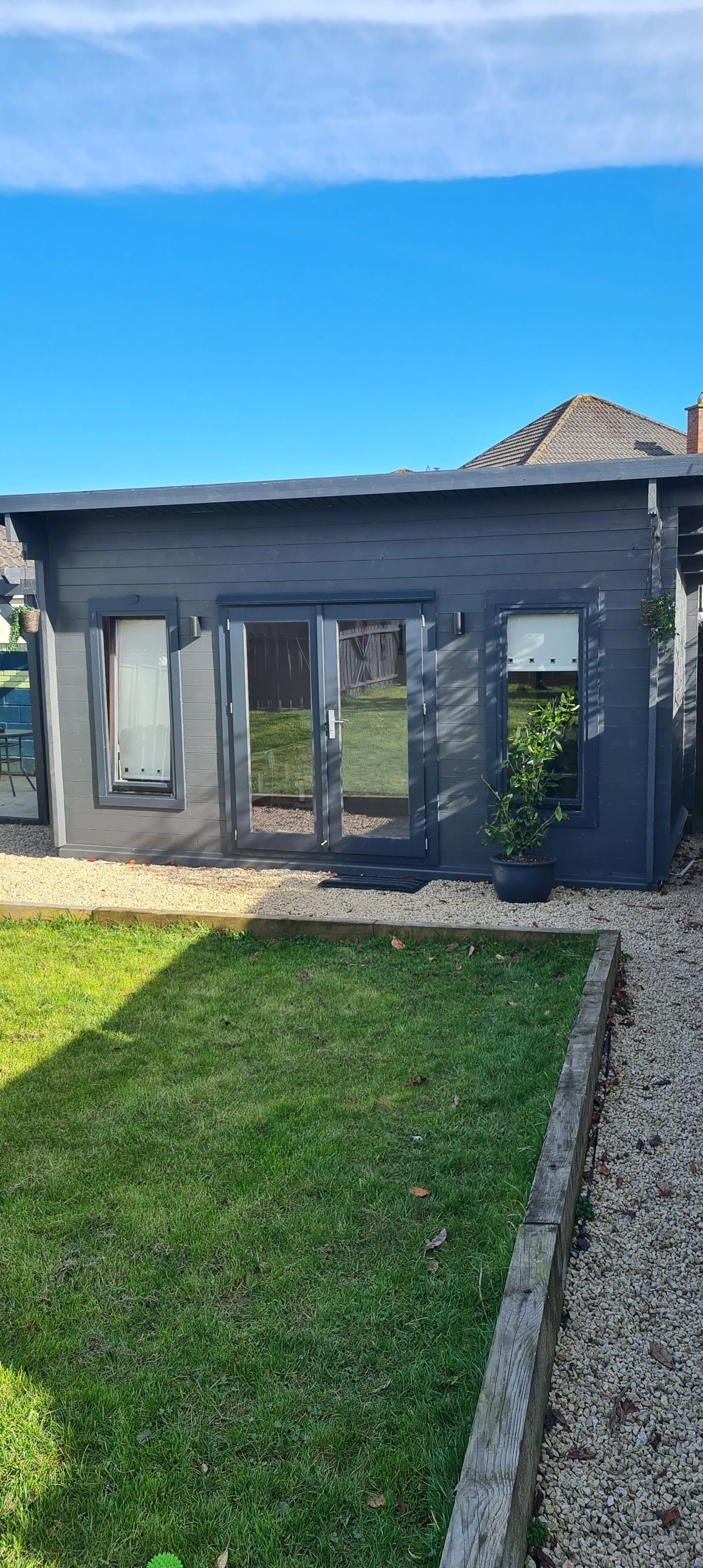 Small, modern dark gray garden office with large windows and glass doors, surrounded by a well-maintained lawn and gravel path under a clear blue sky.