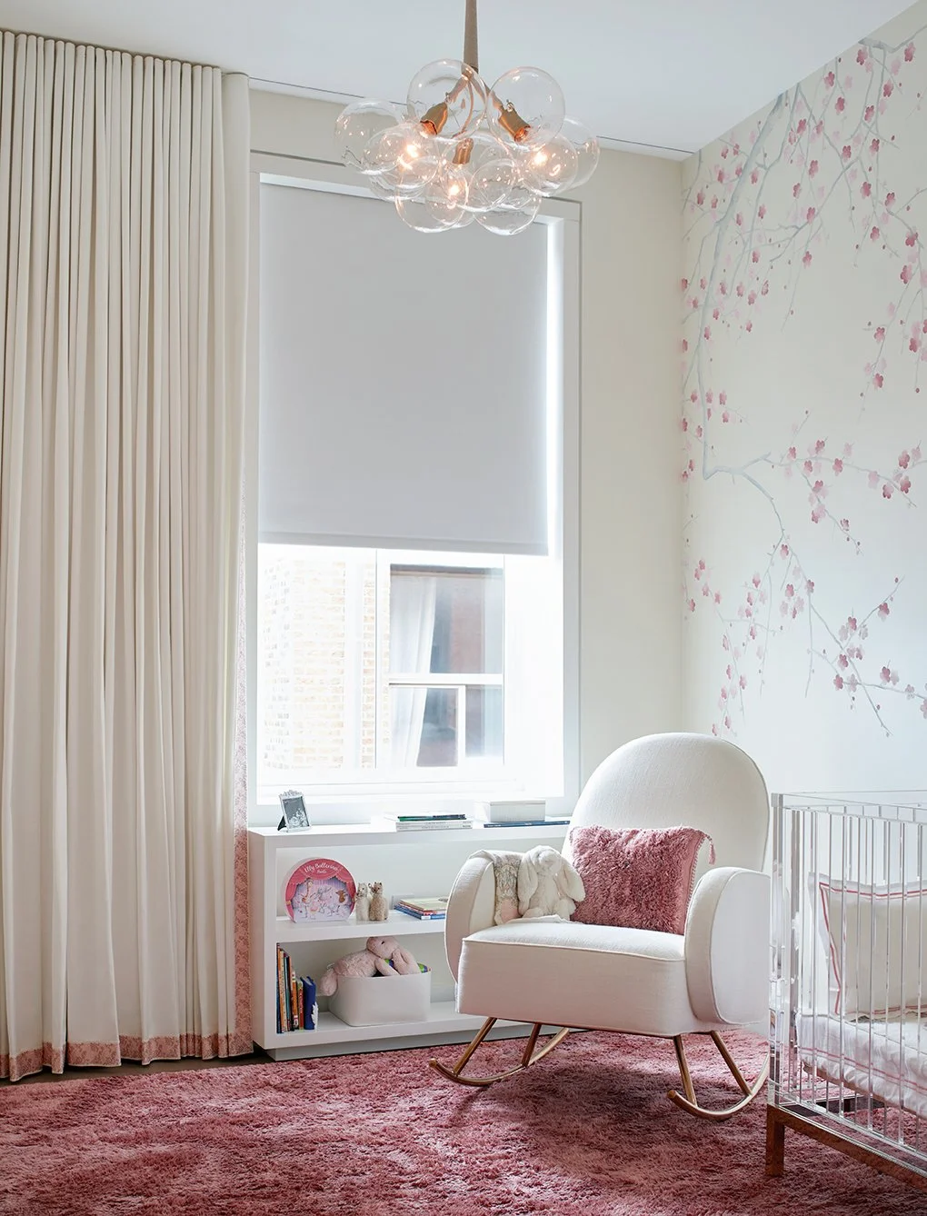 A nursery with a white armchair with a pink pillow and stuffed bunny, a white bookshelf with toys and books, a crib, floral wallpaper, large window with curtains and blinds, and a pink carpet.