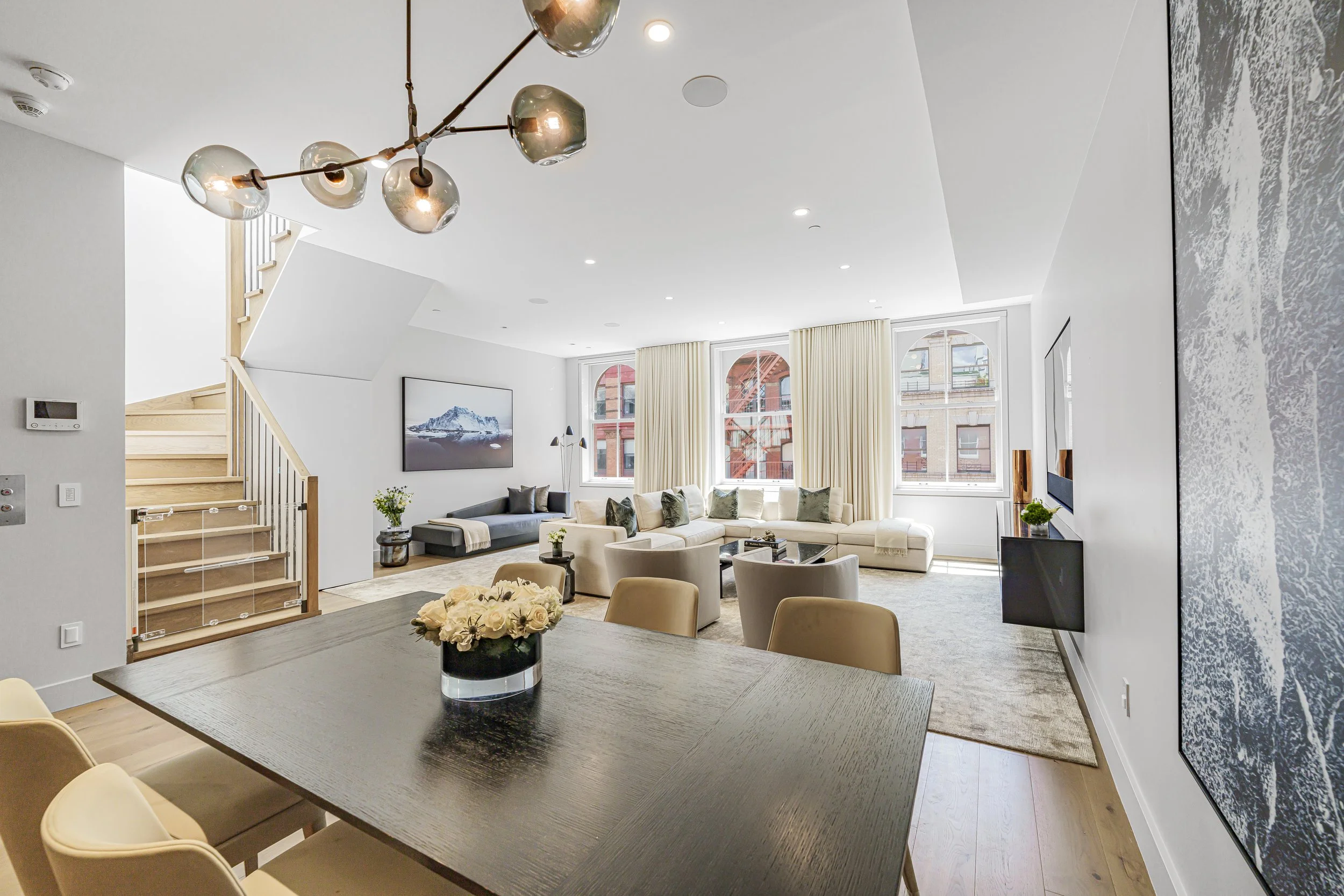 Modern living room with white walls, large windows, beige curtains, a black wall mounted television, a gray sectional sofa, decorative cushions, a coffee table, and an abstract wall art. There is a dining table with a flower arrangement and a stairca