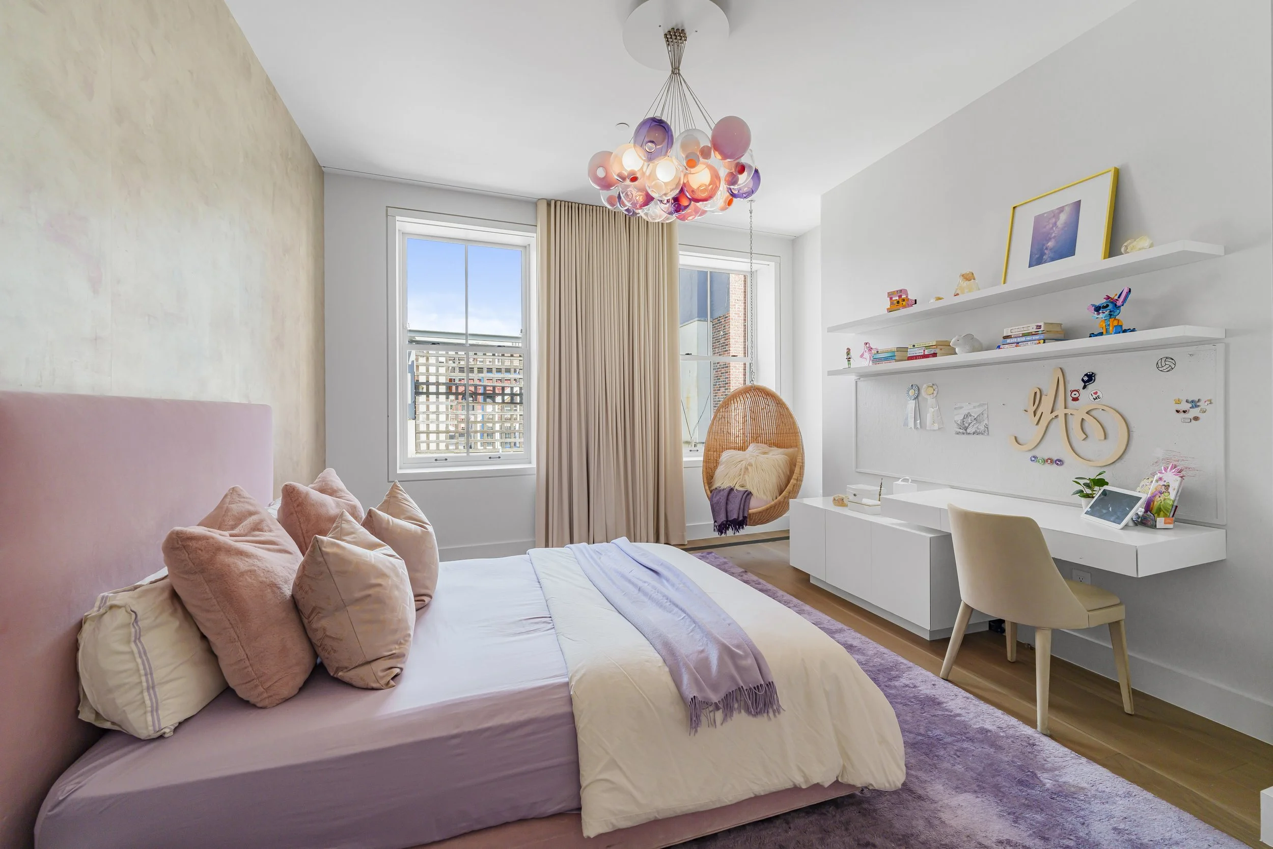Bright bedroom with pink bed and pillows, beige curtains, a hanging basket chair with a pillow and purple throw, white shelves with toys and books, a beige chair at a white desk, a purple rug, and a multicolored bubble chandelier.