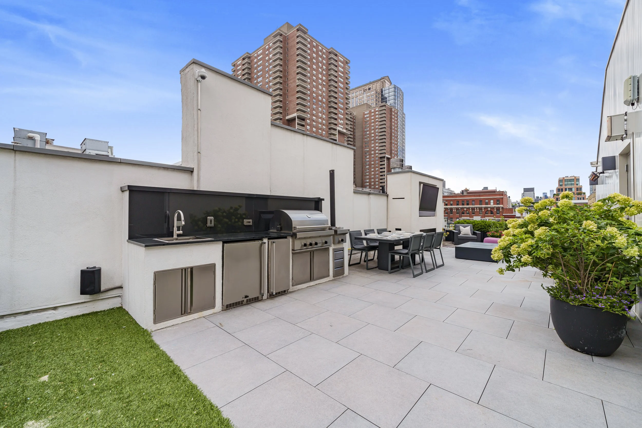 Rooftop patio with outdoor kitchen, dining area, seating lounge, and large potted plant, with city buildings in the background.