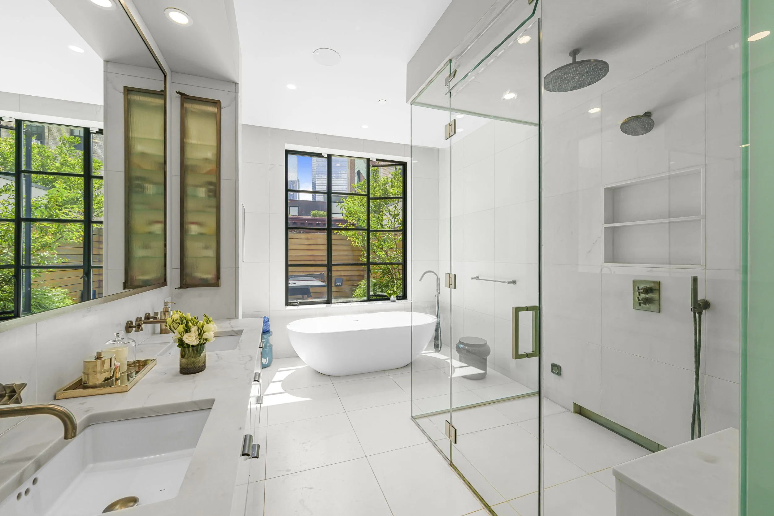 Modern white bathroom with large windows, a freestanding bathtub, glass-enclosed shower, and marble vanity with gold accents.