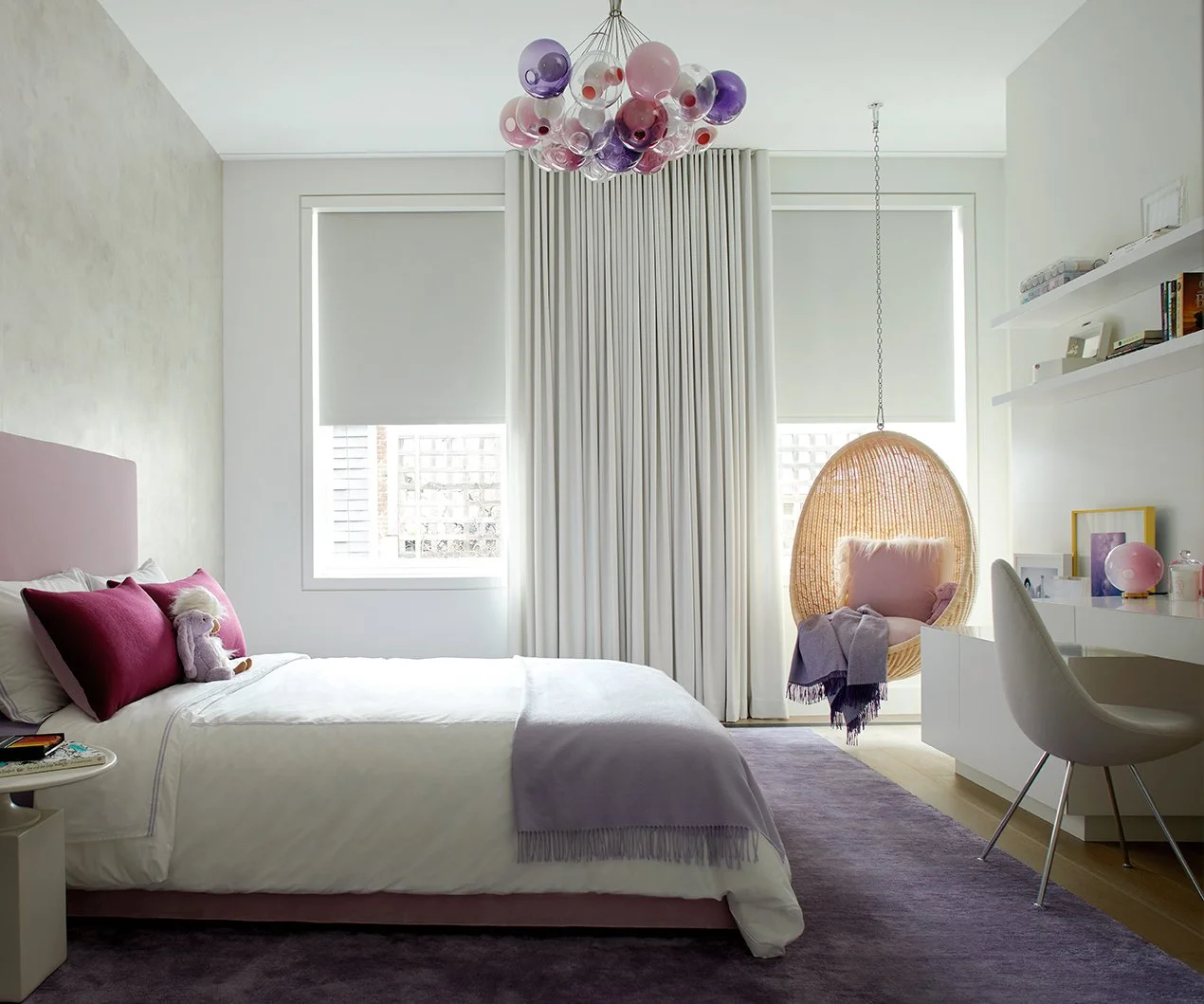 Bright modern bedroom with a white bed, pink pillows, plush toys, a cozy hanging chair with a pink pillow, a lavender blanket, a purple rug, and a white desk with decorative items, large windows with white blinds, and a colorful chandelier.