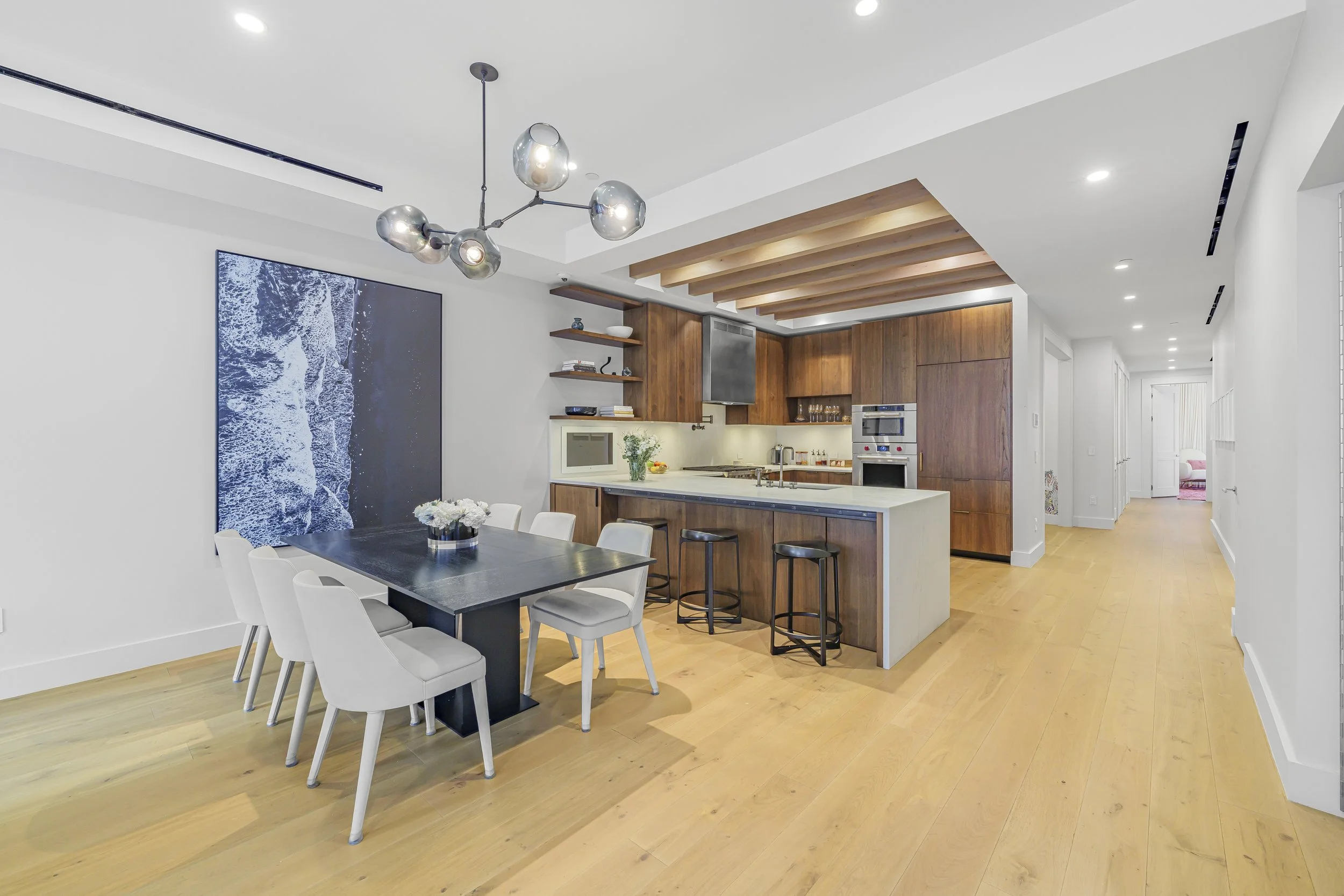 Open-concept kitchen and dining area with white walls, wooden flooring, modern dark dining table with white chairs, a large artwork of ocean waves, wooden kitchen cabinets, and ceiling with recessed lighting and wooden slats.